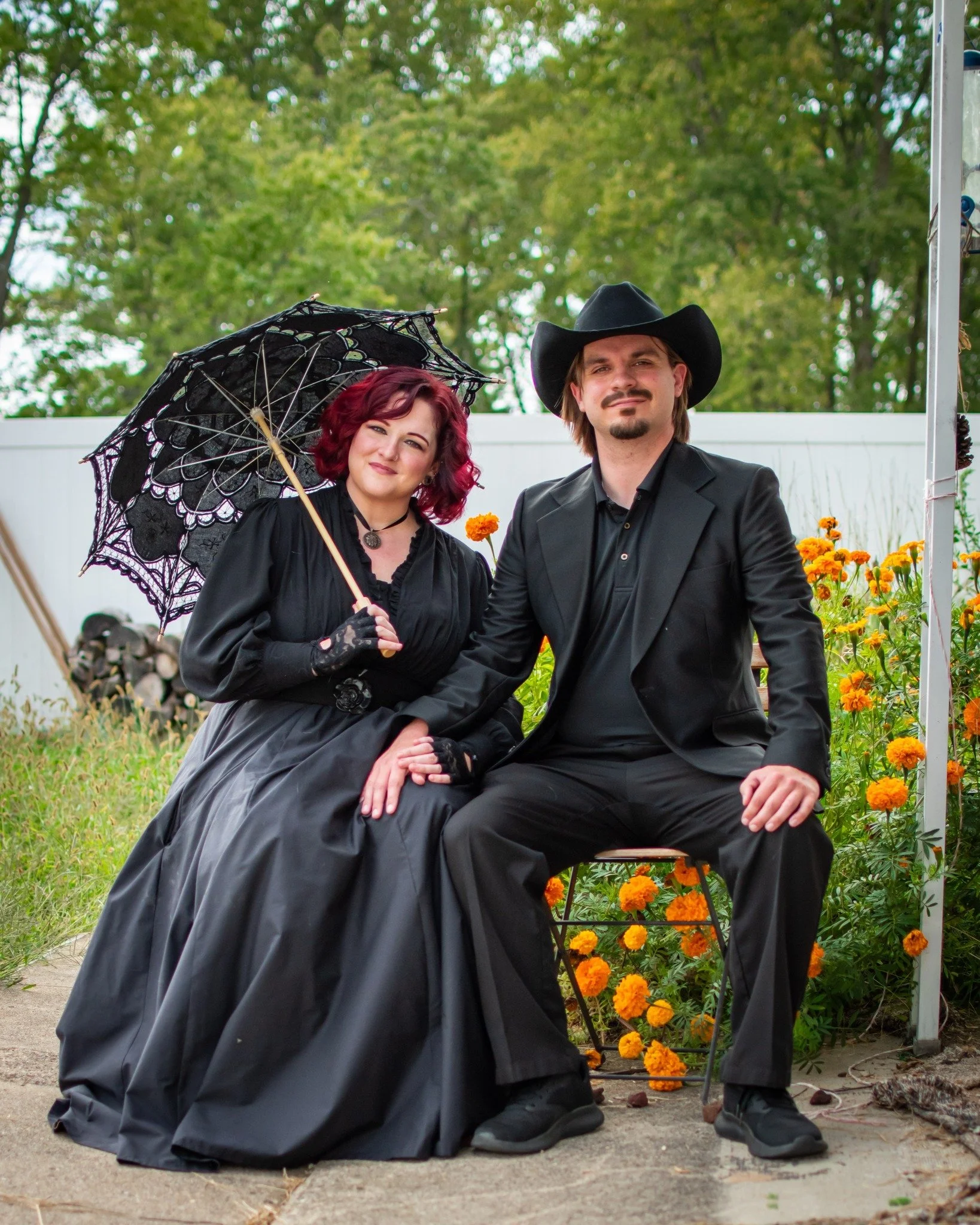 A woman dressed in gothic attire holding a lace parasol and sitting on the ground, posing with a man dressed as a cowboy, sitting on a chair outdoors surrounded by yellow marigolds and greenery.