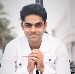 Portrait of a young man with dark hair, wearing a white jacket, smiling with hands clasped in front, outdoors