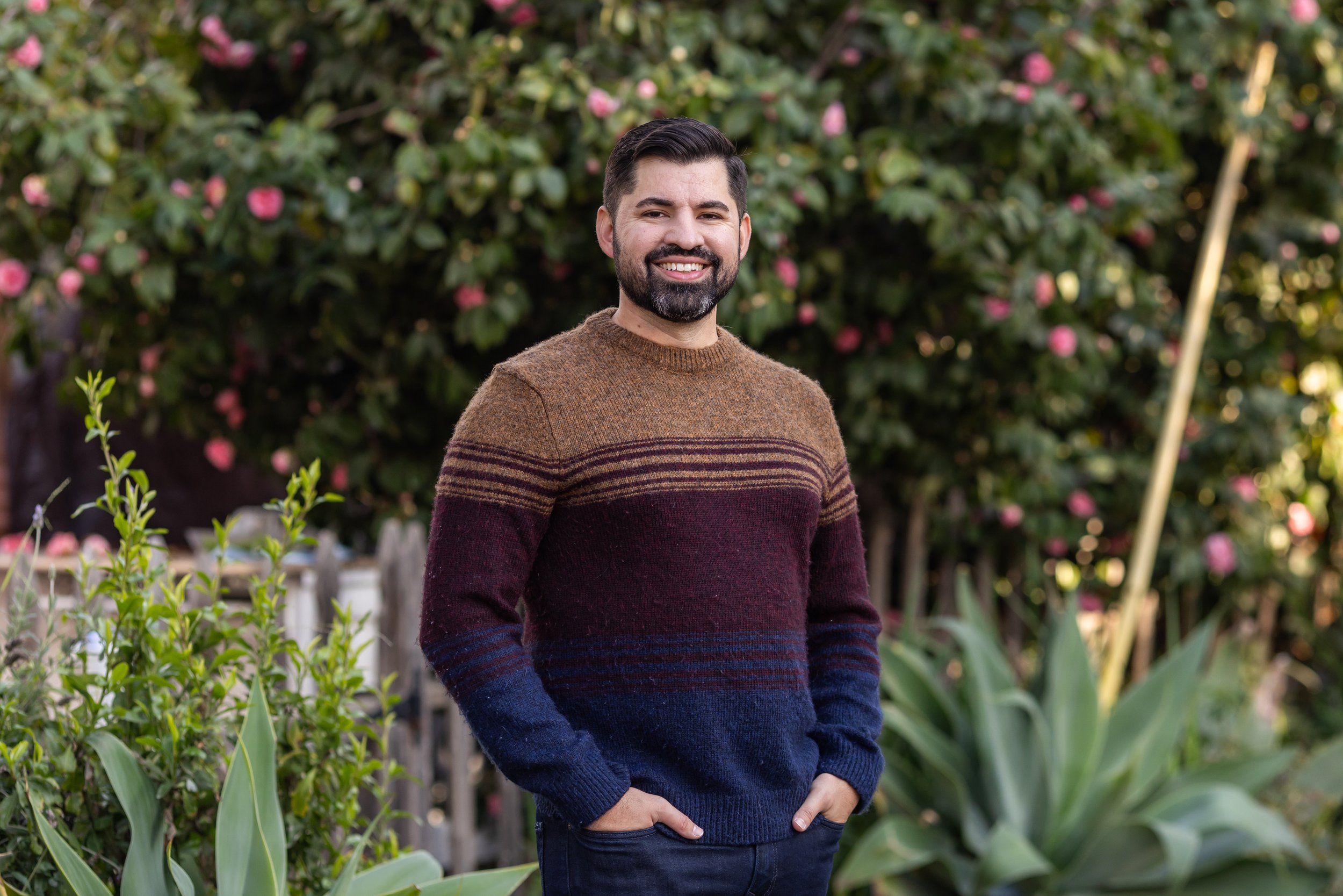 Smiling man with dark hair and beard standing outdoors in front of flowering bushes, wearing a multicolored sweater and blue jeans.