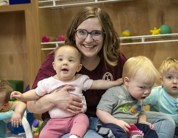 Smiling daycare teacher holding a baby girl while sitting on the floor with three young children in a child care classroom.