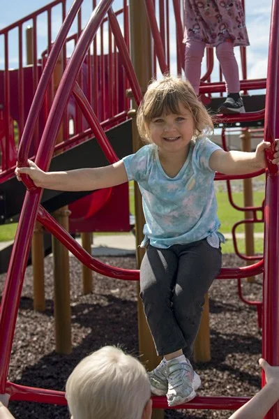 A young girl from Grandkids child care smiling while standing on a red spiral climbing structure at a playground, with other children nearby.