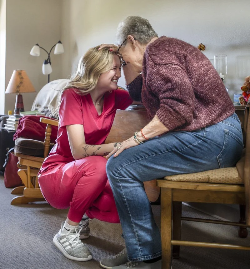A young nurse and an resident of The Maples assisted living are smiling and touching foreheads in a joyful moment inside a home.