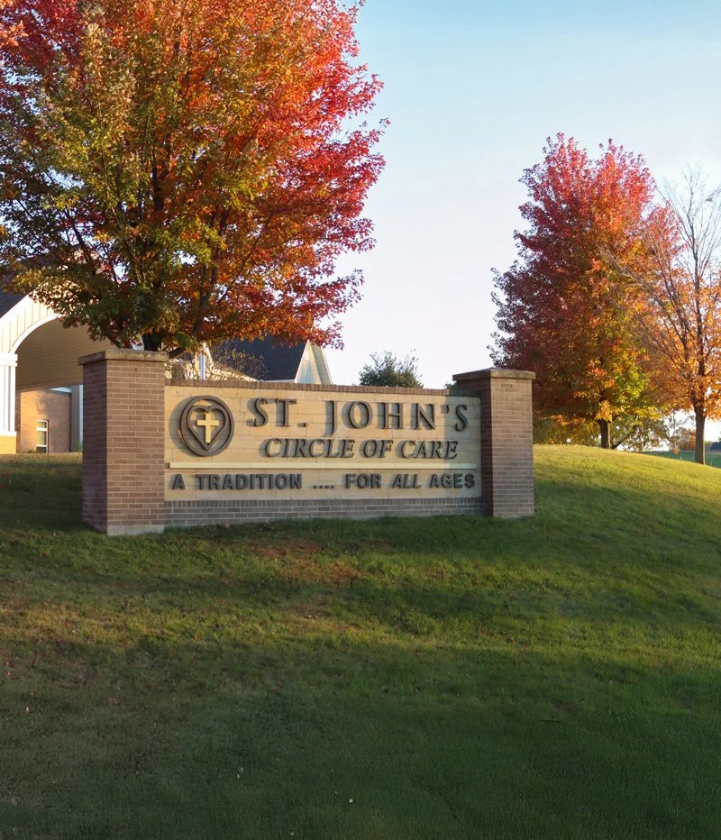 Sign for St. John's Circle of Care, with a cross symbol, surrounded by trees with autumn leaves, on a grassy hill.
