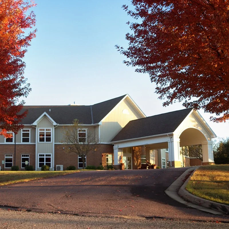 Exterior of The Maples assisted living covered entrance, white and brick exterior, surrounded by trees with autumn foliage, and a clear blue sky above.