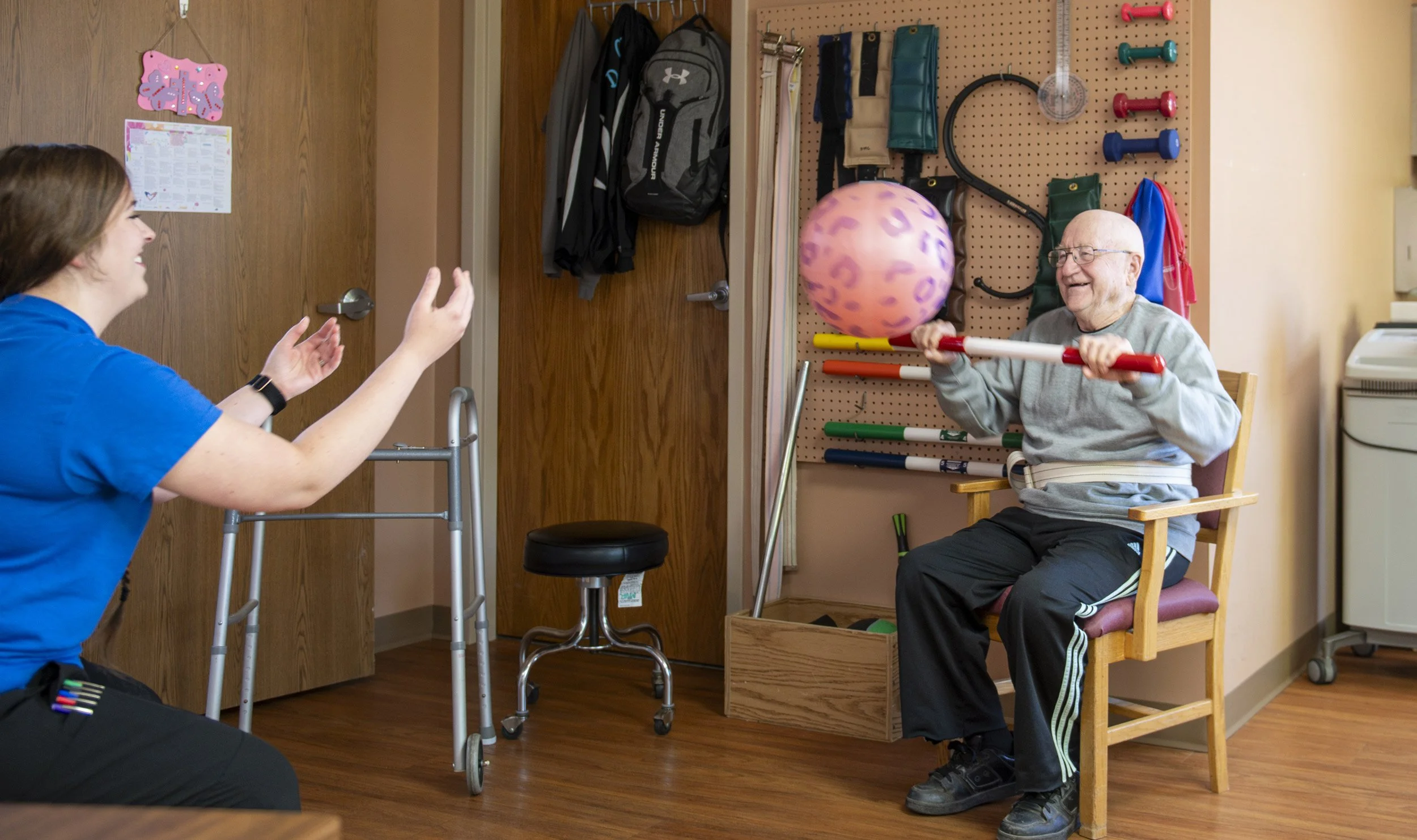 An elderly man with glasses, sitting on a wooden chair, playing with a therapy ball suspended in the air, while a physical therapist interacts with him in a therapy room.