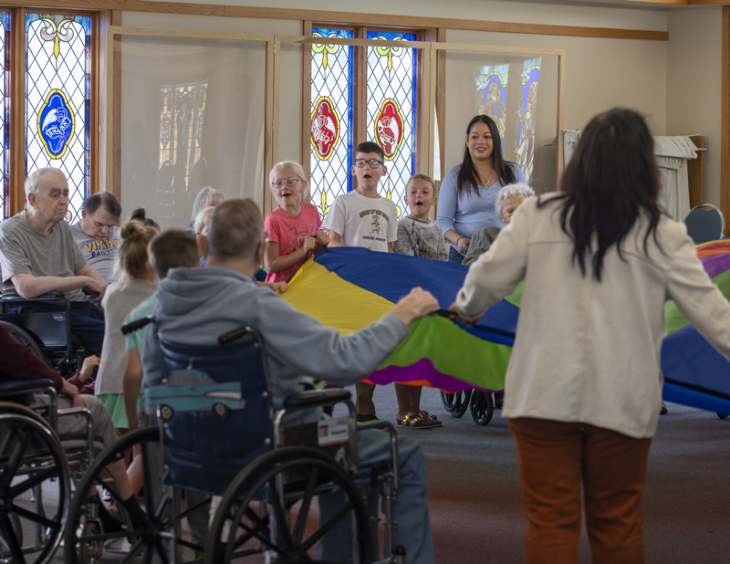 Children, residents, employees, and volunteers participate in a group activity with a multicolored parachute in the Chapel of the Good Shepherd in Springfield MN