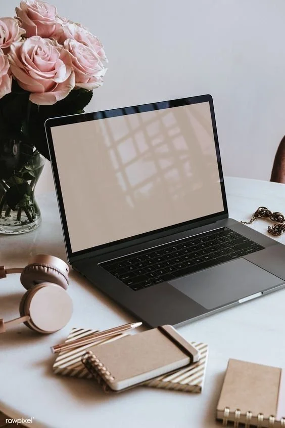A laptop on a white desk with a bouquet of pink roses in a glass vase, a pair of beige headphones, a gold pen, and several notebooks.
