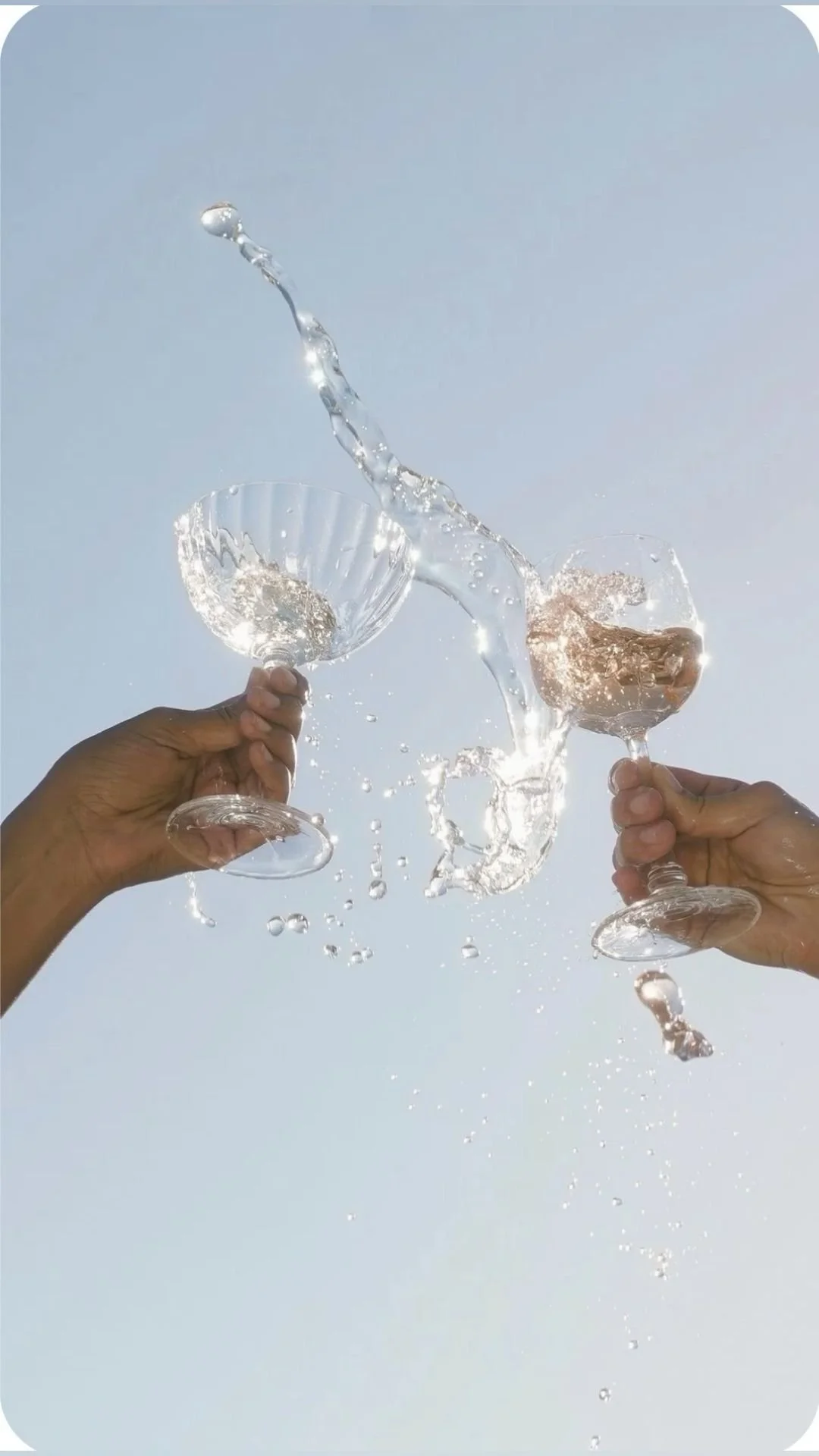 Two hands holding champagne glasses clinking together with water splashing as they drop, against a clear sky background.