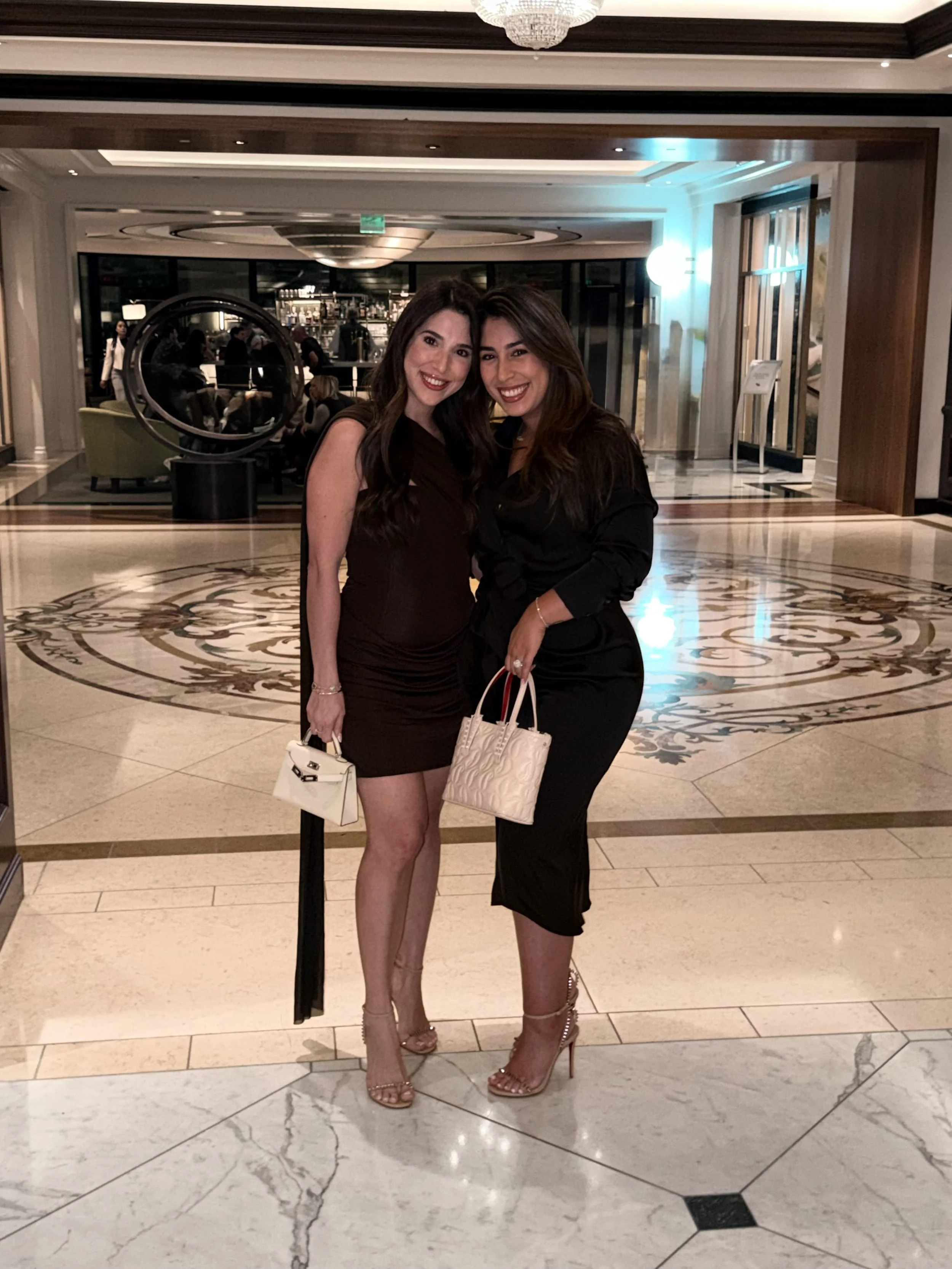 Two women in black dresses and heels posing together in a hotel lobby, smiling, holding white handbags.