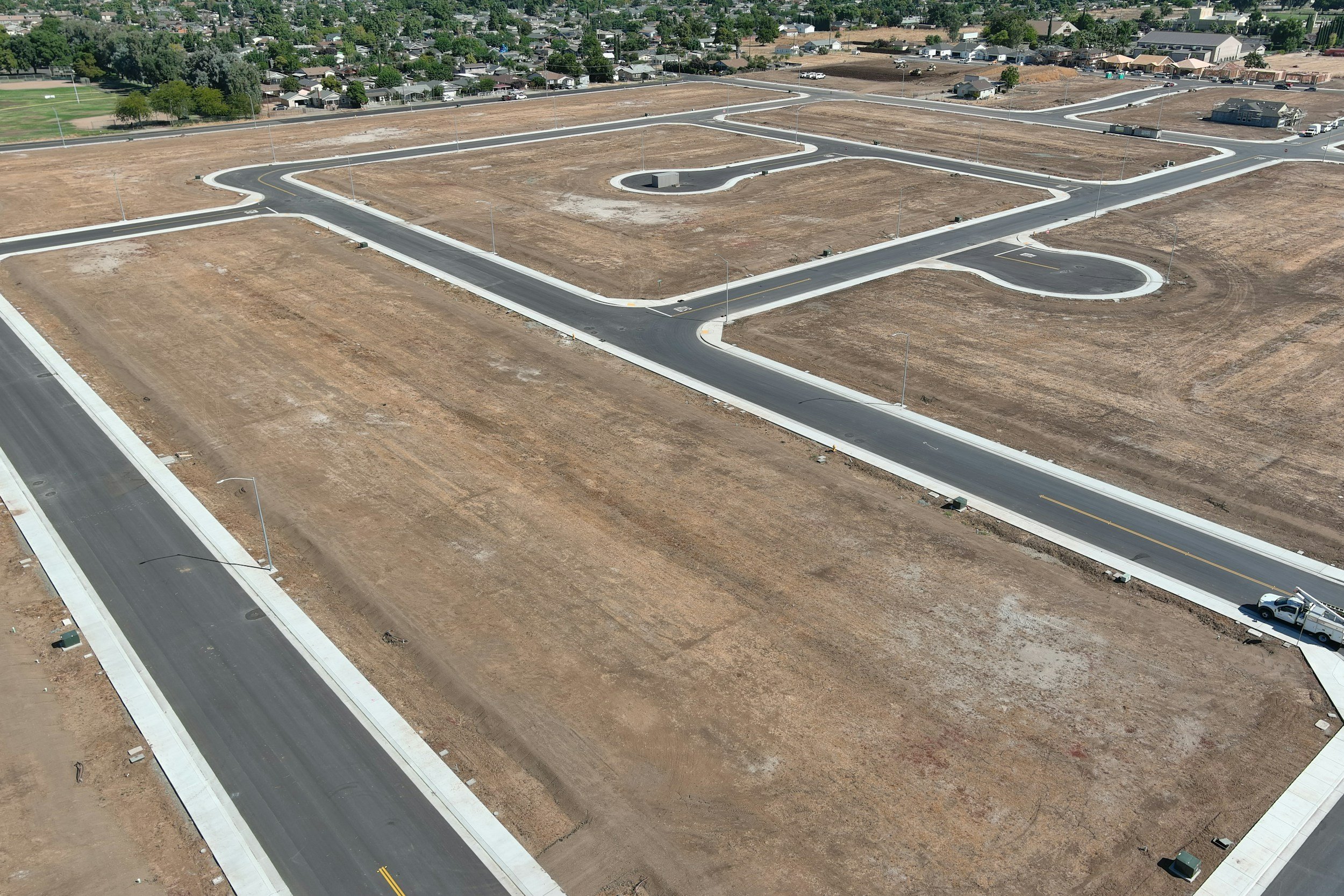 Aerial view of a developing residential area with newly paved roads, streetlights, and empty plots of land for future homes, with a few houses in the background.