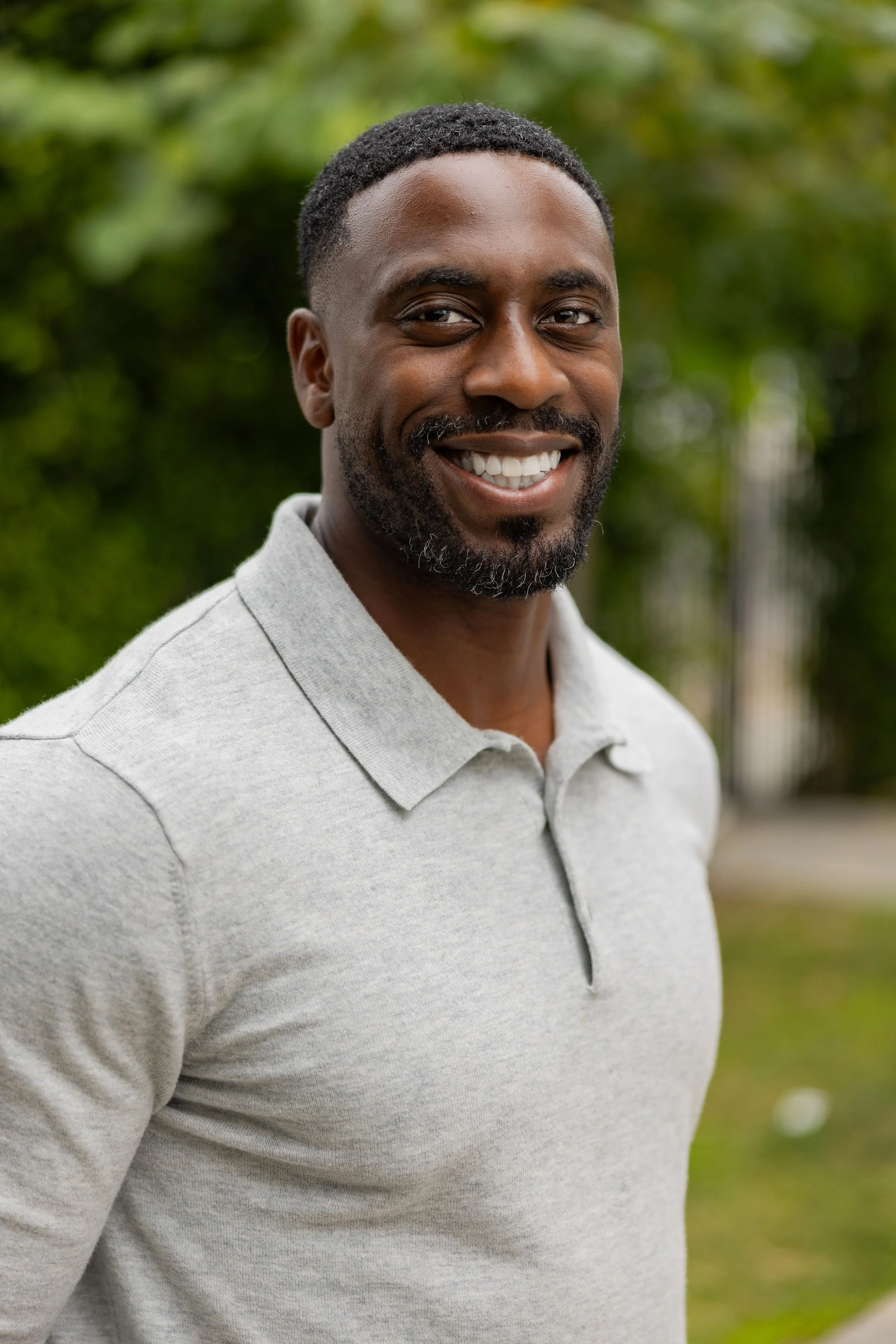 A smiling man with short black hair and a beard, wearing a light gray polo shirt outdoors with blurred green foliage in the background.