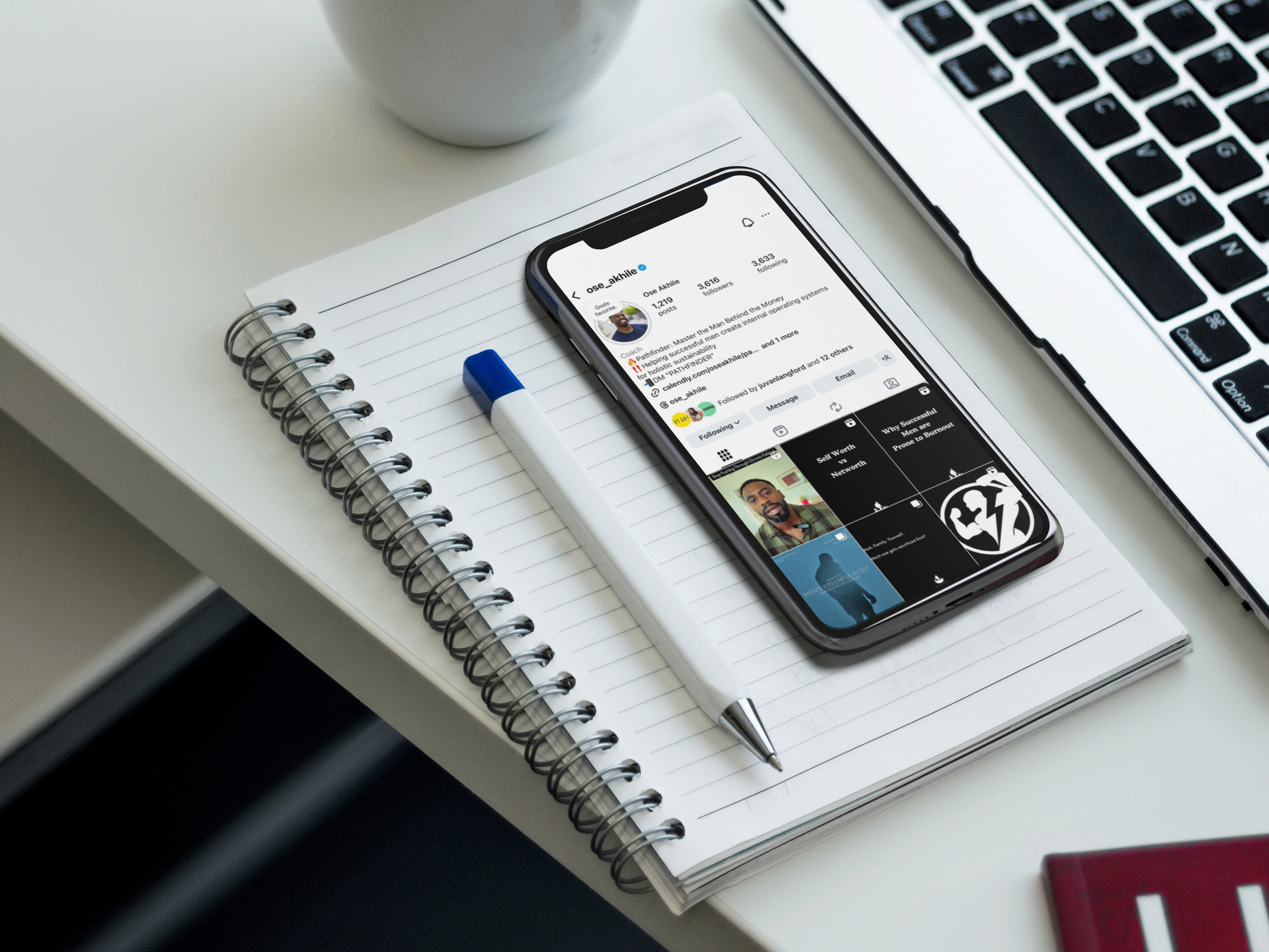 Close-up of a white desk with an open notebook, a white and blue pen, a smartphone displaying a social media profile, a laptop, and a white cup.