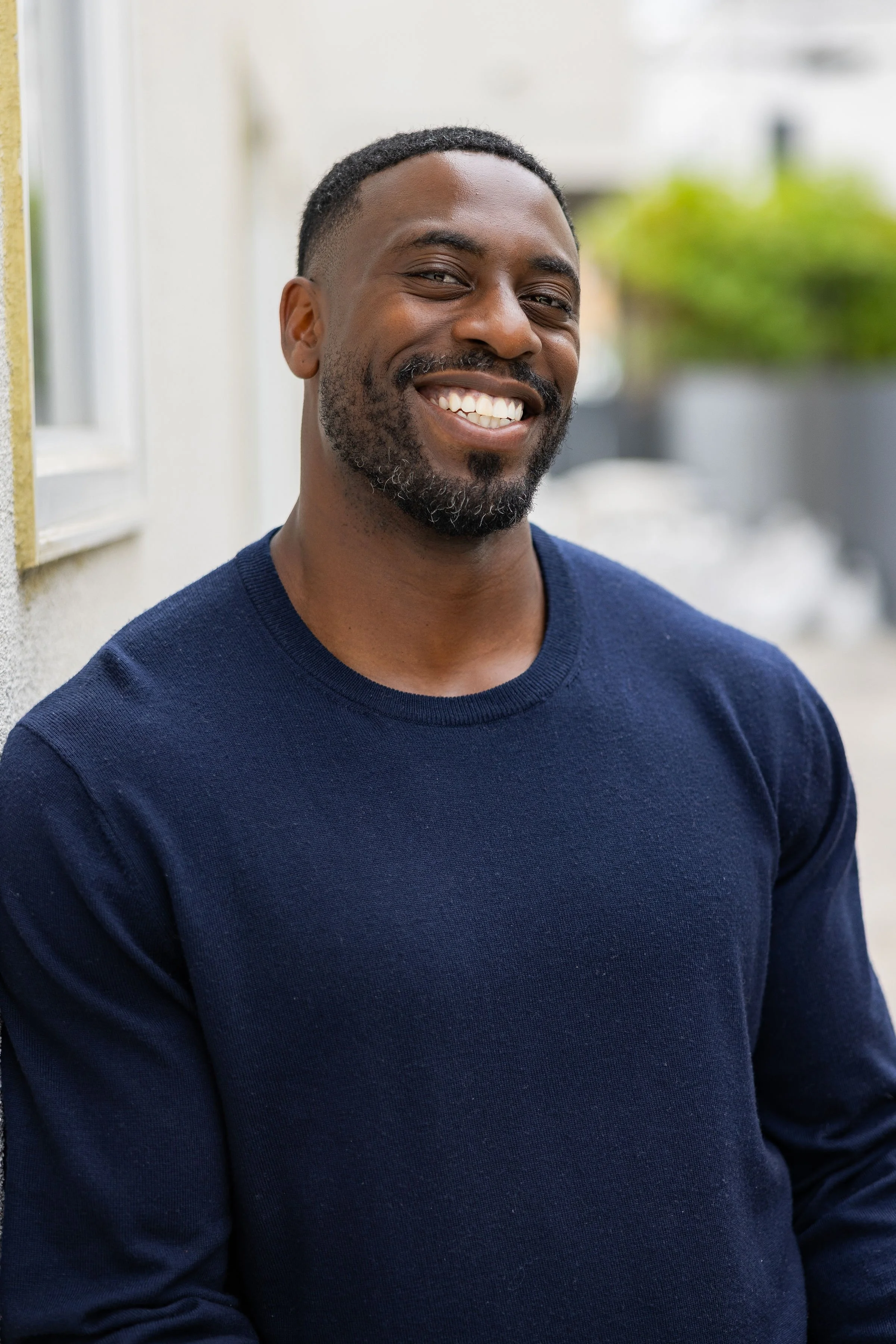 A smiling man with a beard, wearing a navy blue sweater, standing outdoors near a building with blurred greenery in the background.
