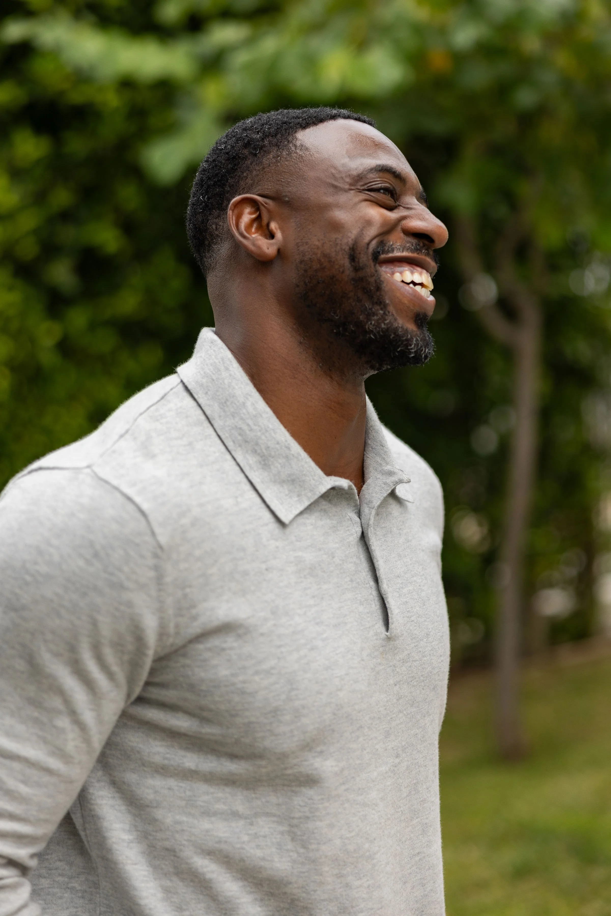 A man outdoors smiling and looking to his right, wearing a light gray polo shirt, with trees and greenery in the background.