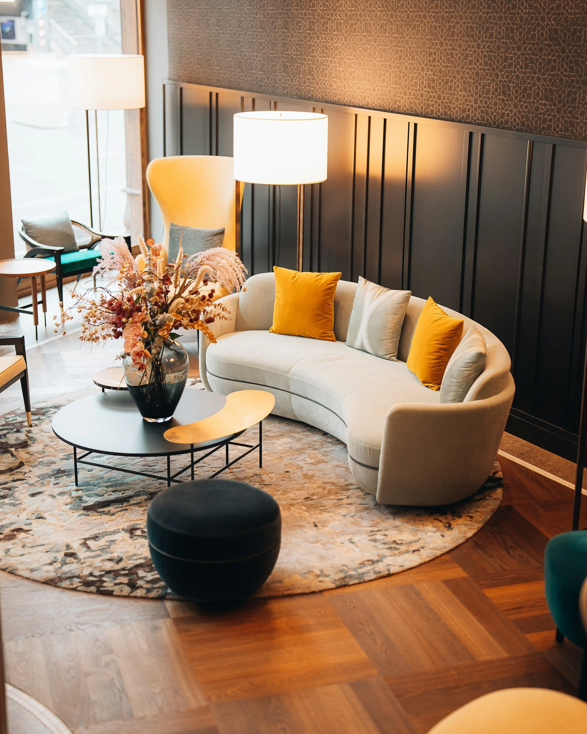 A stylish hotel lobby with a curved cream-colored sofa, yellow and light blue accent pillows, and a round black coffee table with a floral arrangement. There are two white floor lamps, a beige armchair, and large windows letting in natural light.