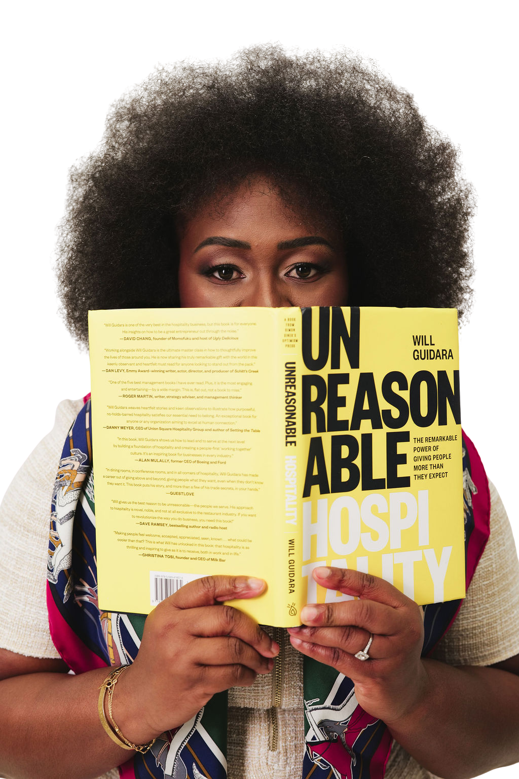 A woman with natural curly hair and makeup, holding a yellow book titled "Unreasonable Hospitality" by Will Guidara, partially obscuring her face.