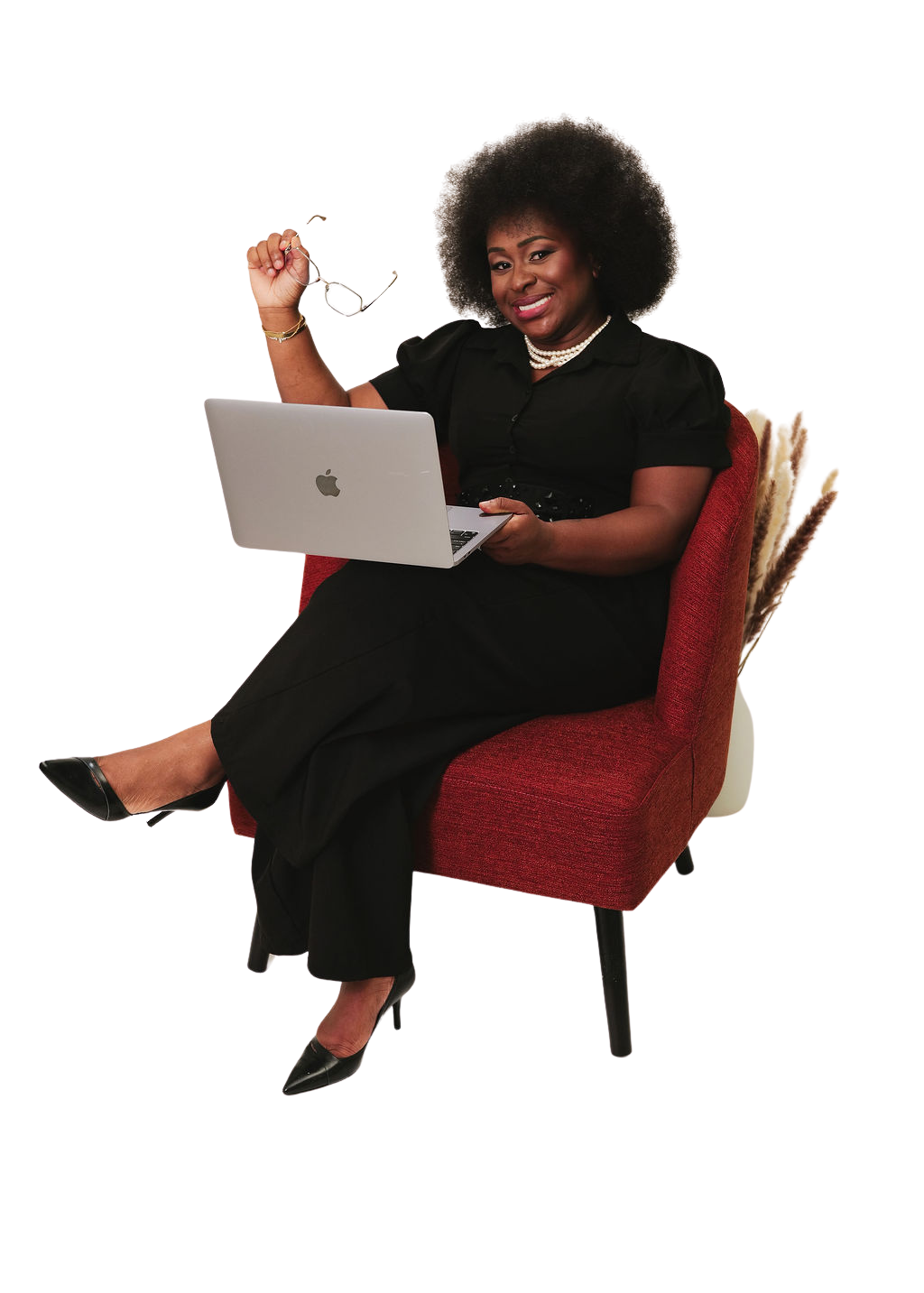 A smiling woman with an afro hairstyle is sitting in a red chair, holding a laptop in her lap and showing a small digital device in her right hand. She is dressed in a black outfit with pearl jewelry.