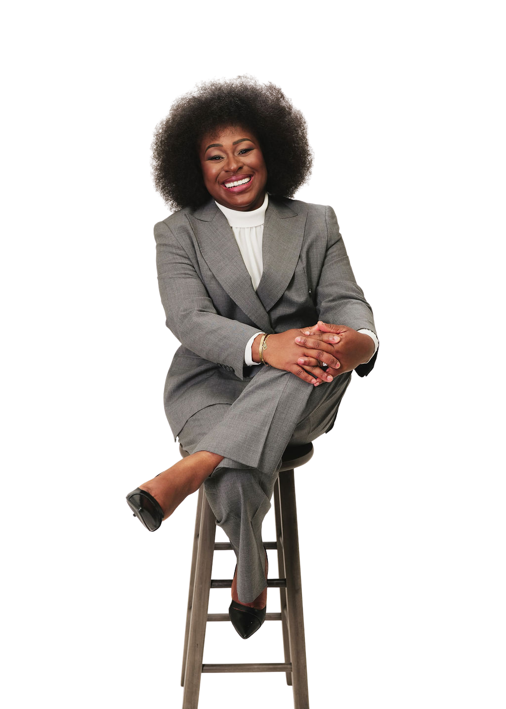 A smiling African American woman in a gray business suit sitting on a stool with one leg crossed over the other, arms crossed on her knee, wearing black heels and a white blouse, against a plain background.