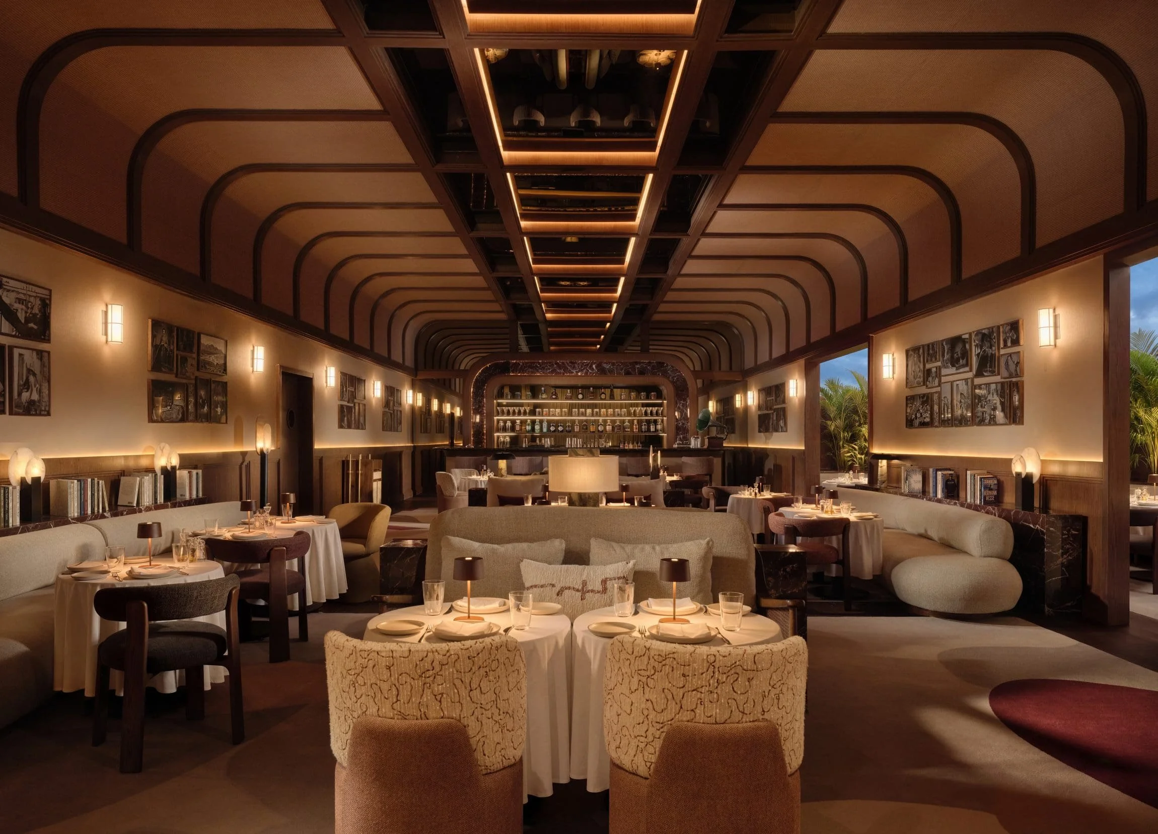 Elegant dimly lit restaurant interior with curved wooden ceiling beams, framed black-and-white photos on walls, and round tables set with plates, glasses, and small lamps.