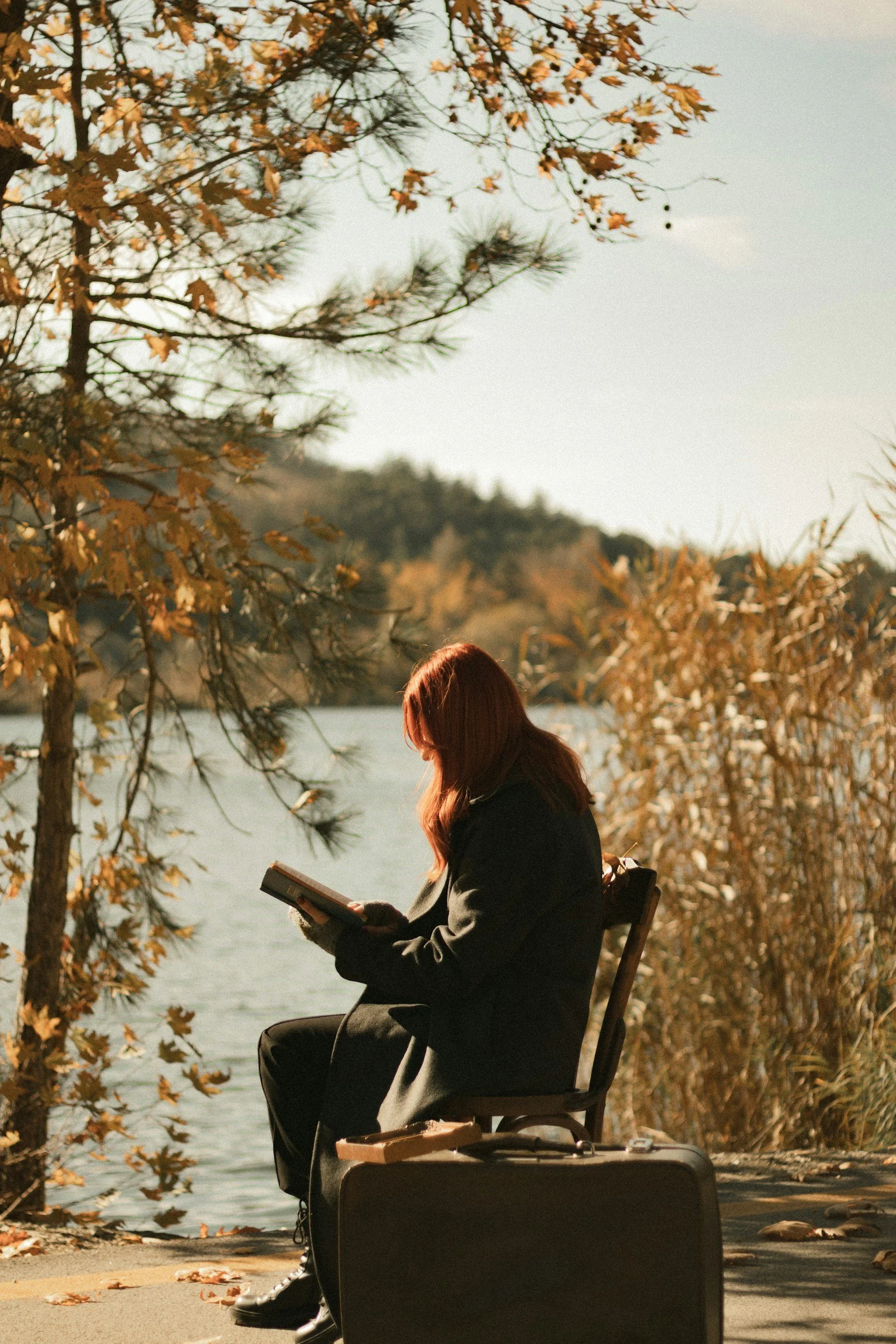 A woman with reddish-brown hair sitting on a bench near a lake reading a book, with a suitcase nearby, surrounded by fall foliage.
