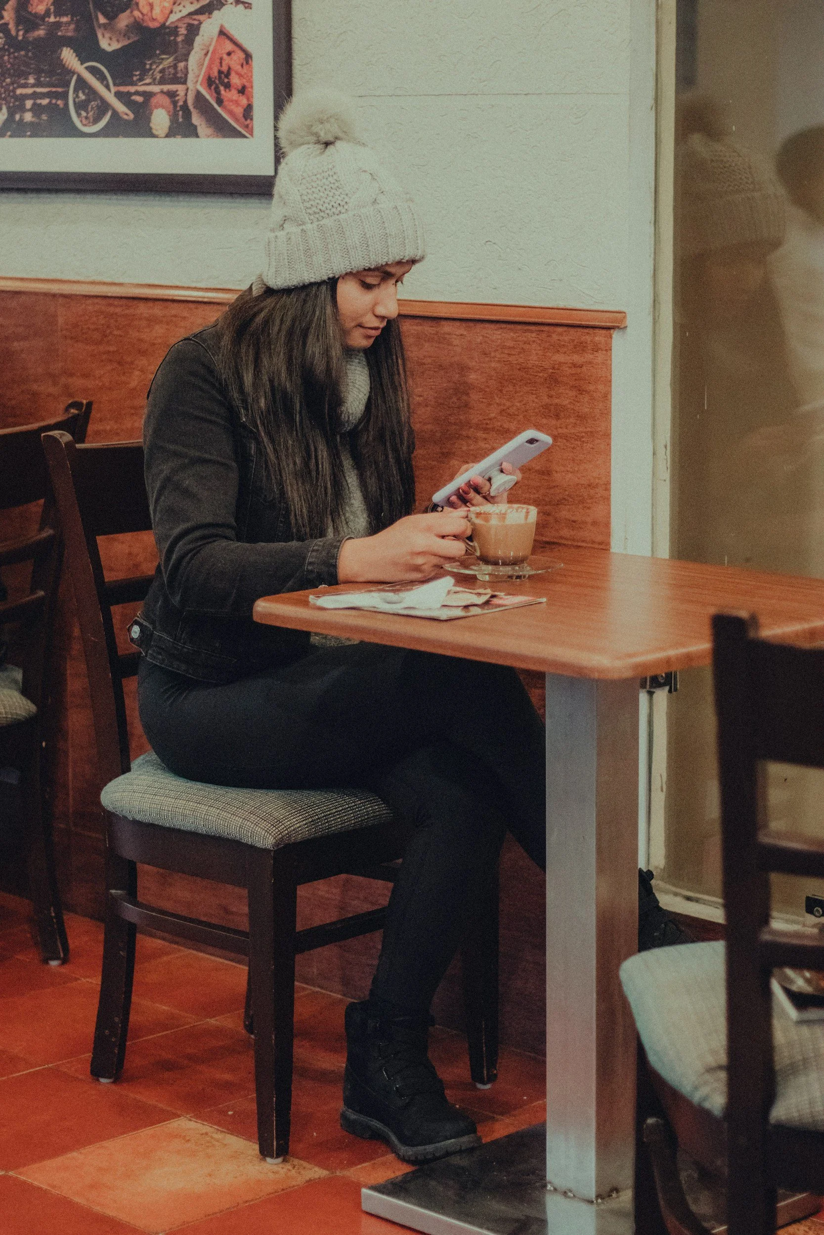 A young woman sitting at a table in a coffee shop, looking at her smartphone, with a cup of coffee in front of her.