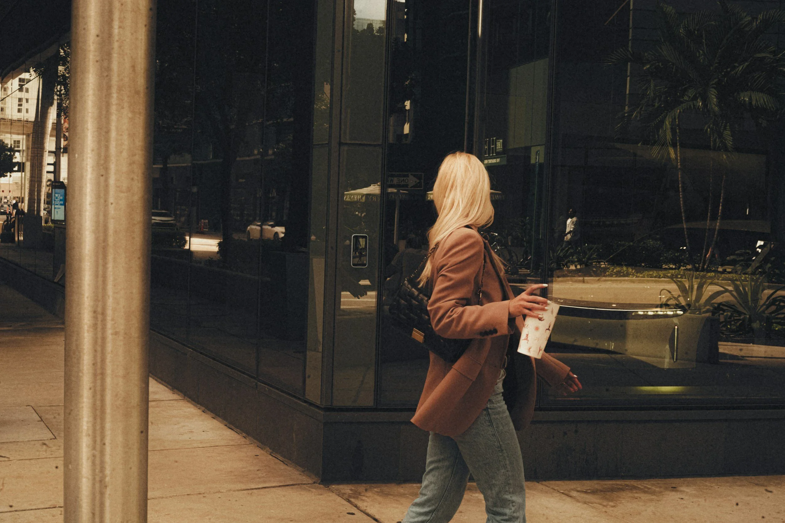 A woman with blonde hair, wearing a brown blazer and blue jeans, walking past a large glass building in an urban area, holding a drink and ignoring her phone, reflected in the window.
