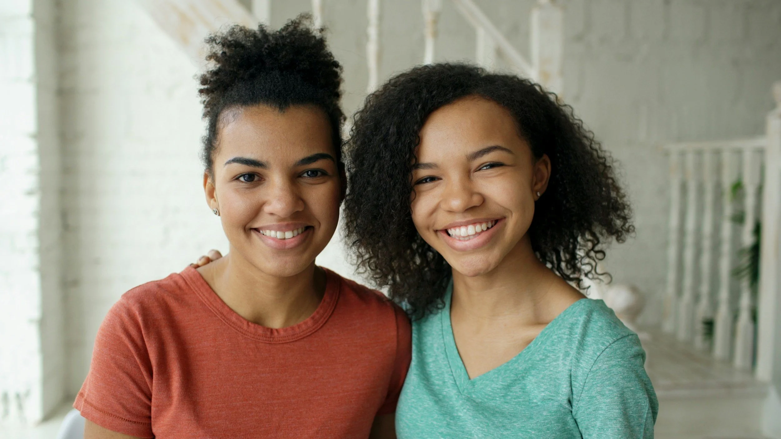 Two women with curly hair smiling and standing close together in a bright indoor setting.