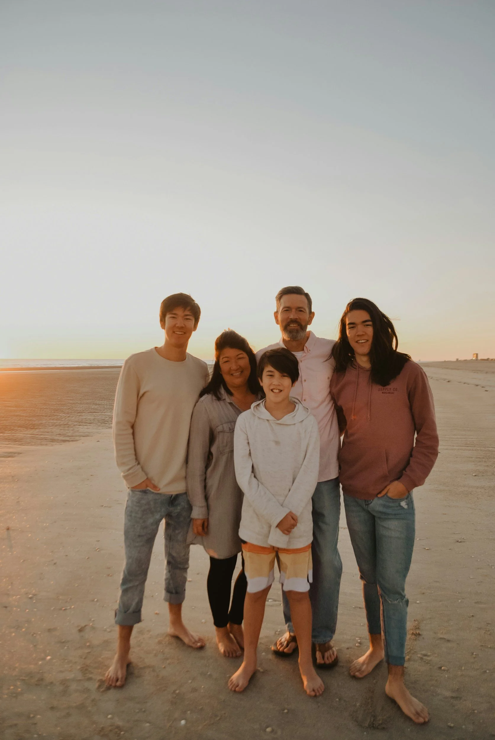 A group of six people standing on a beach at sunset, smiling and posing for a photo. The background shows the ocean and a clear sky.