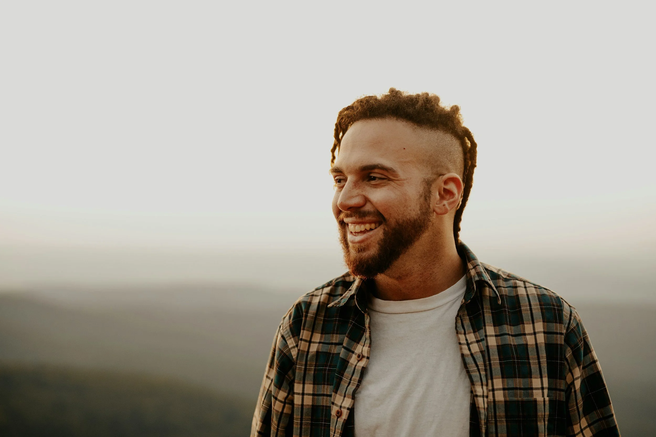 A smiling man with a beard wearing a plaid shirt and white t-shirt, outdoors at sunset or sunrise, with a blurred natural background.