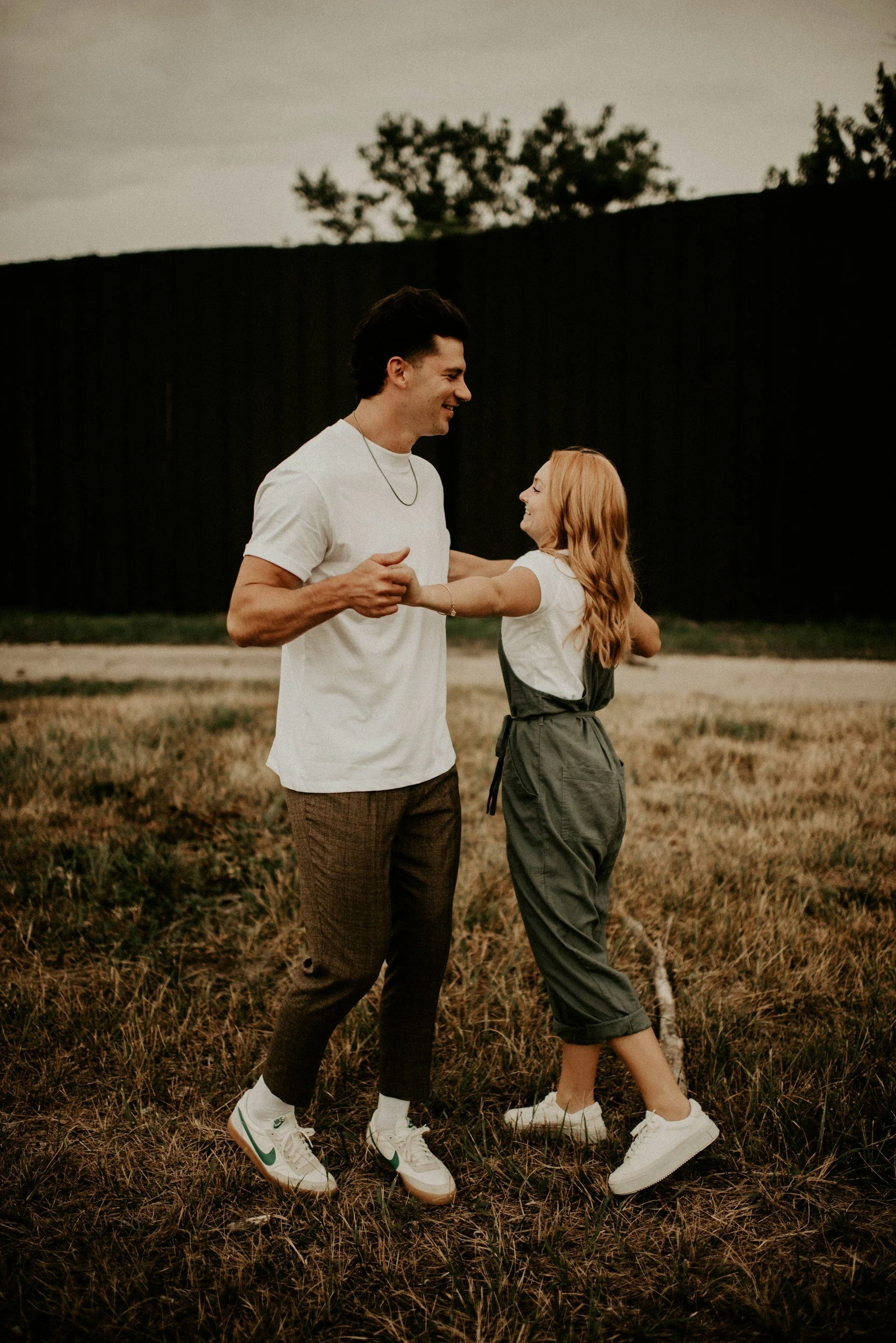 A young man and girl, holding hands and smiling, dancing in a field at dusk. They are wearing casual clothing and sneakers, with a dark fence and trees in the background.