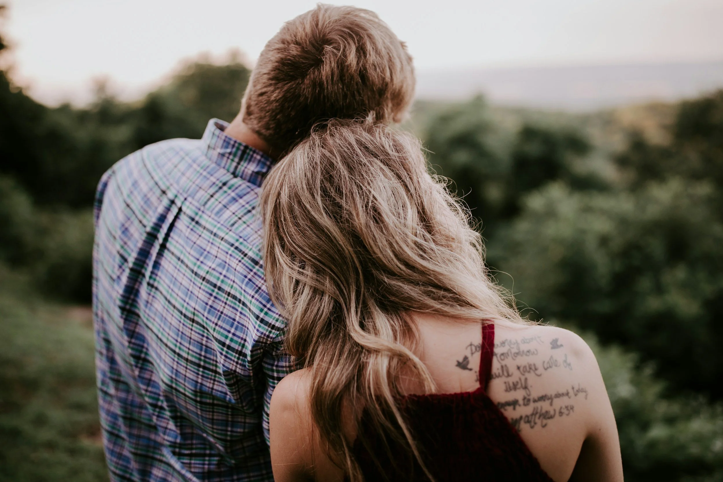 A couple embracing outdoors, the man has light brown hair and is wearing a plaid shirt, the woman has long wavy hair and a tattoo on her upper arm, wearing a sleeveless dress with writing on it.