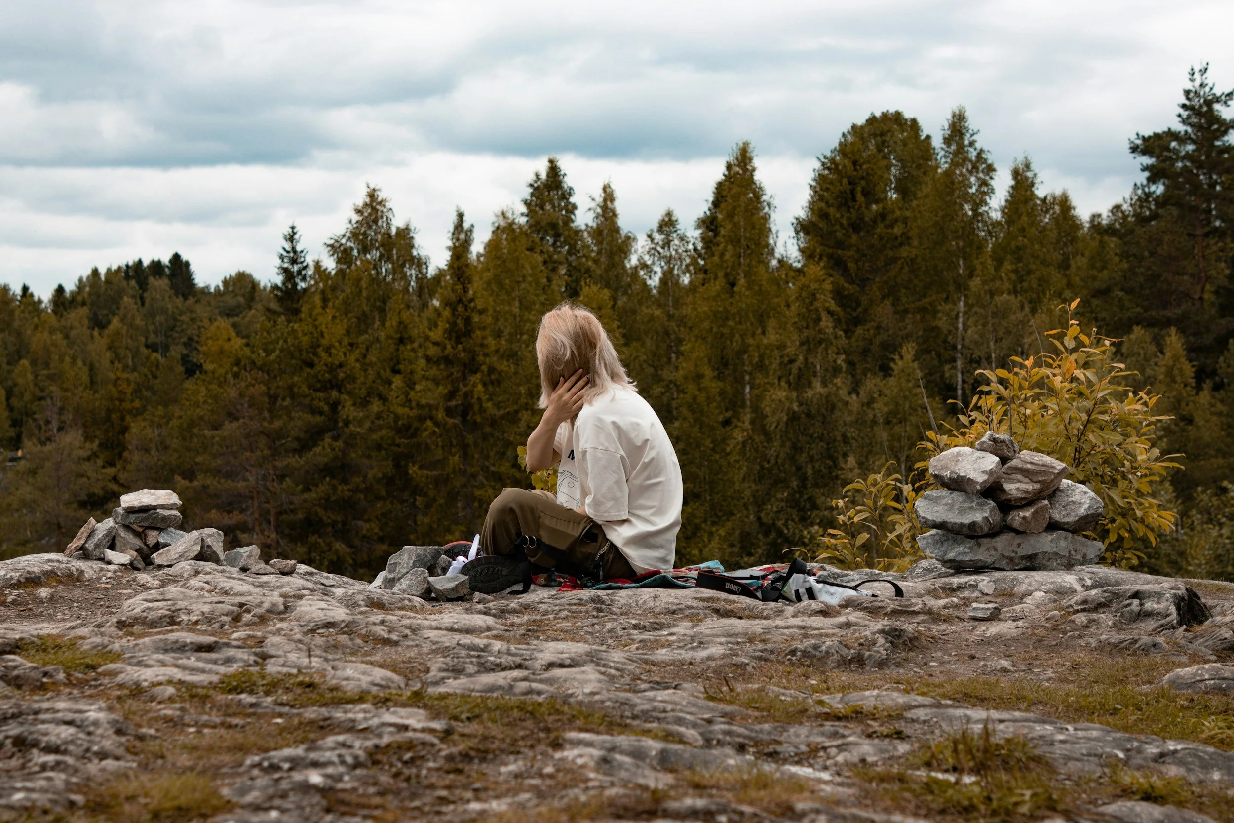 Person with blonde hair sitting on rocky ground next to a small pile of stones, with a forest of evergreen trees in the background and cloudy sky overhead.