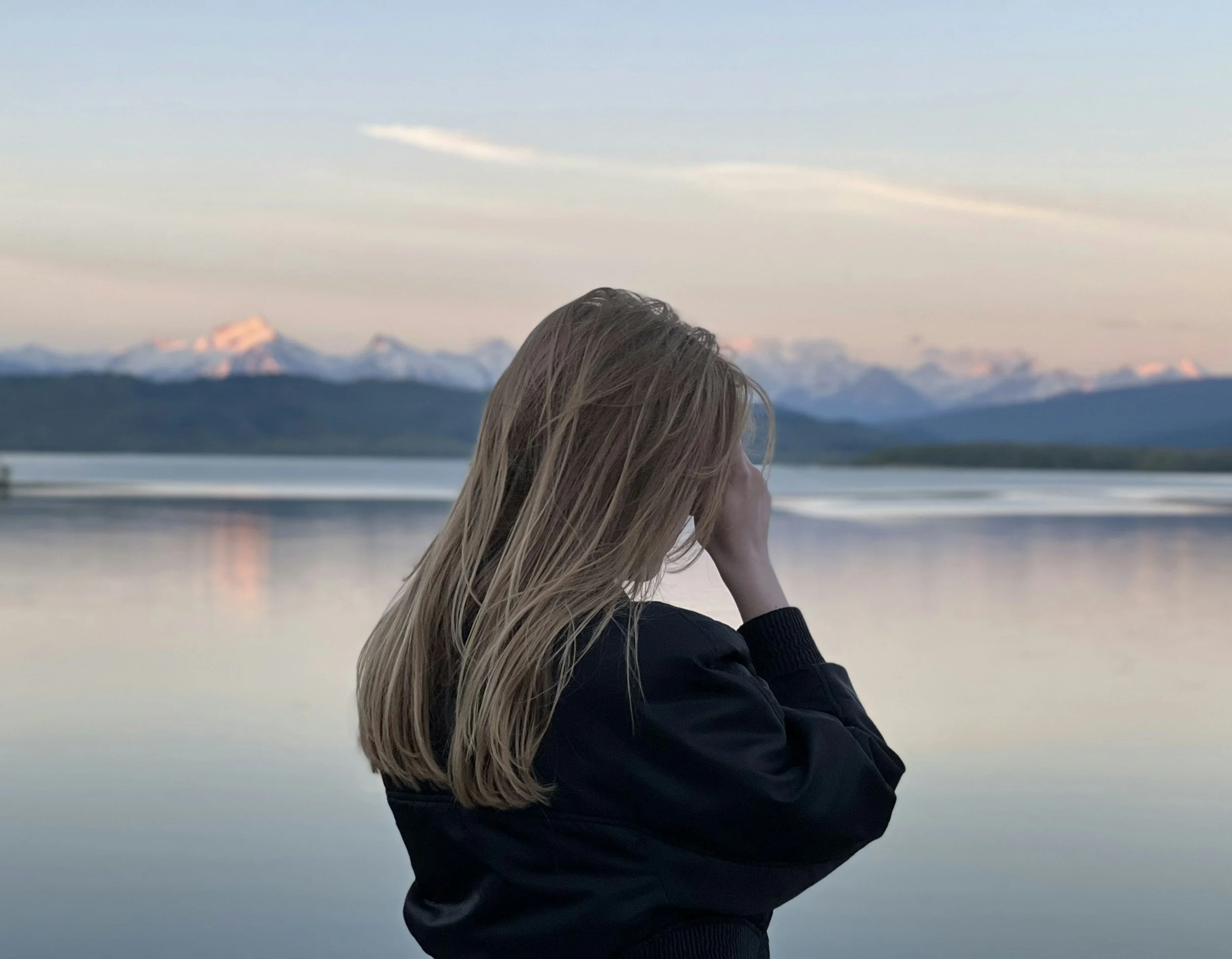 A woman with long blonde hair sitting by a body of water, with snow-capped mountains in the background during sunset.