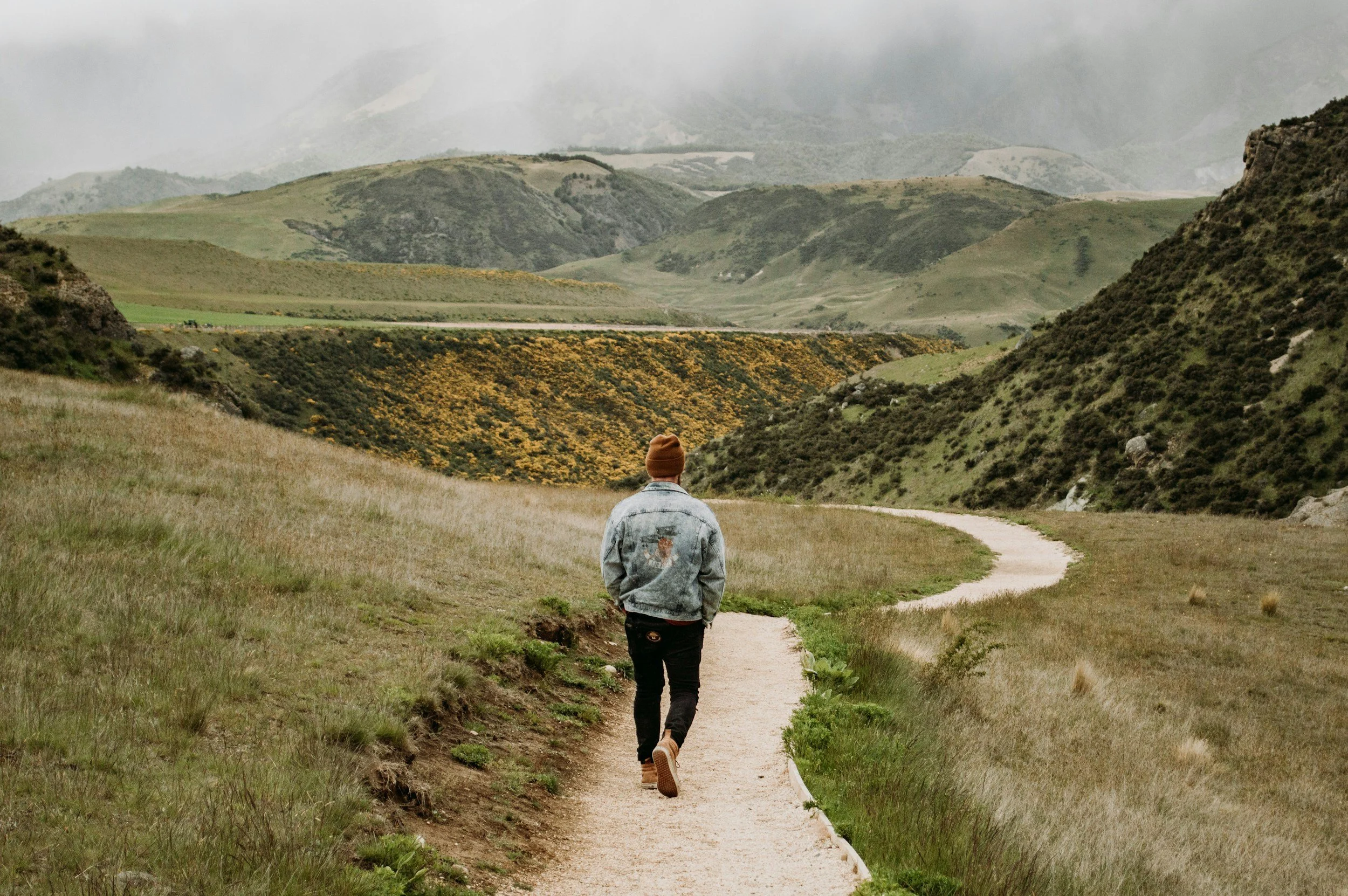 Person walking along a winding dirt trail through grassy hills and mountains under cloudy skies.