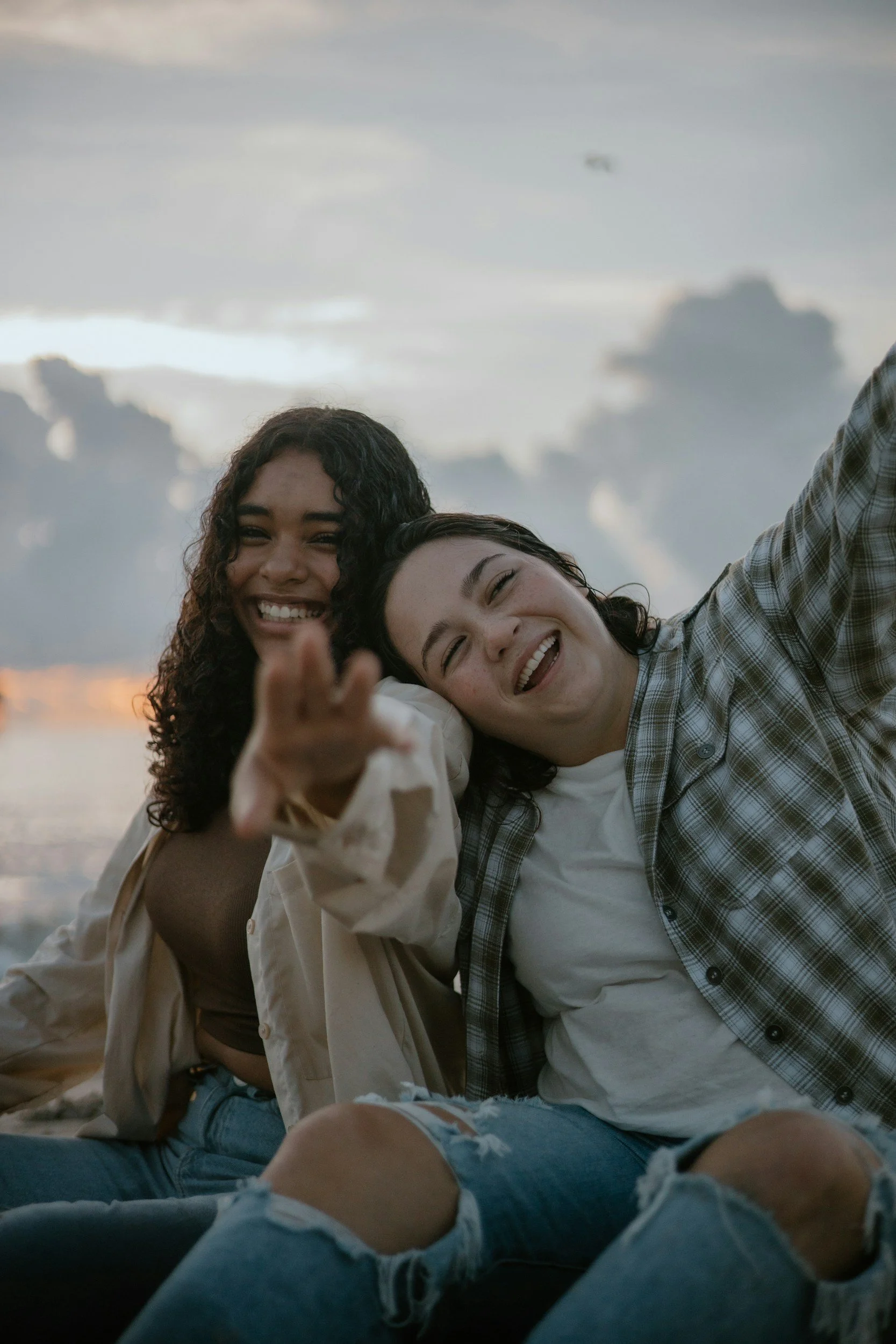 Two women smiling and sitting on a beach with cloudy sky, one wearing a plaid shirt, the other a beige jacket, and both wearing ripped jeans.