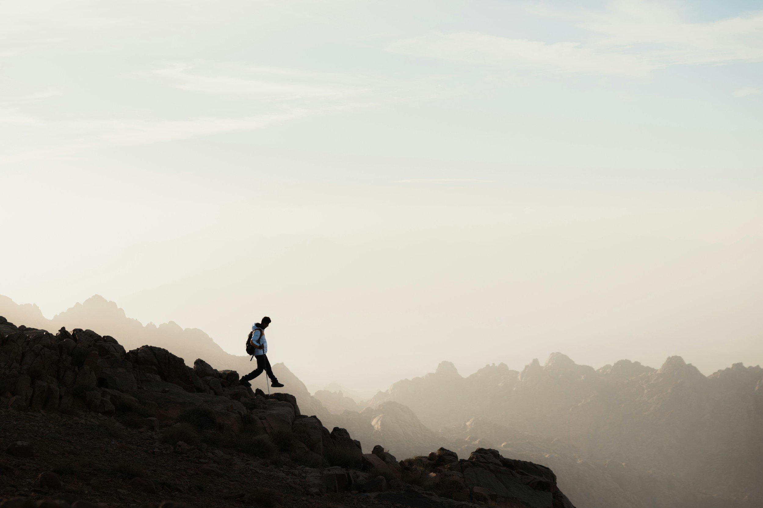 A person hiking on rocky terrain during dawn or dusk with mountains and cloudy sky in the background.