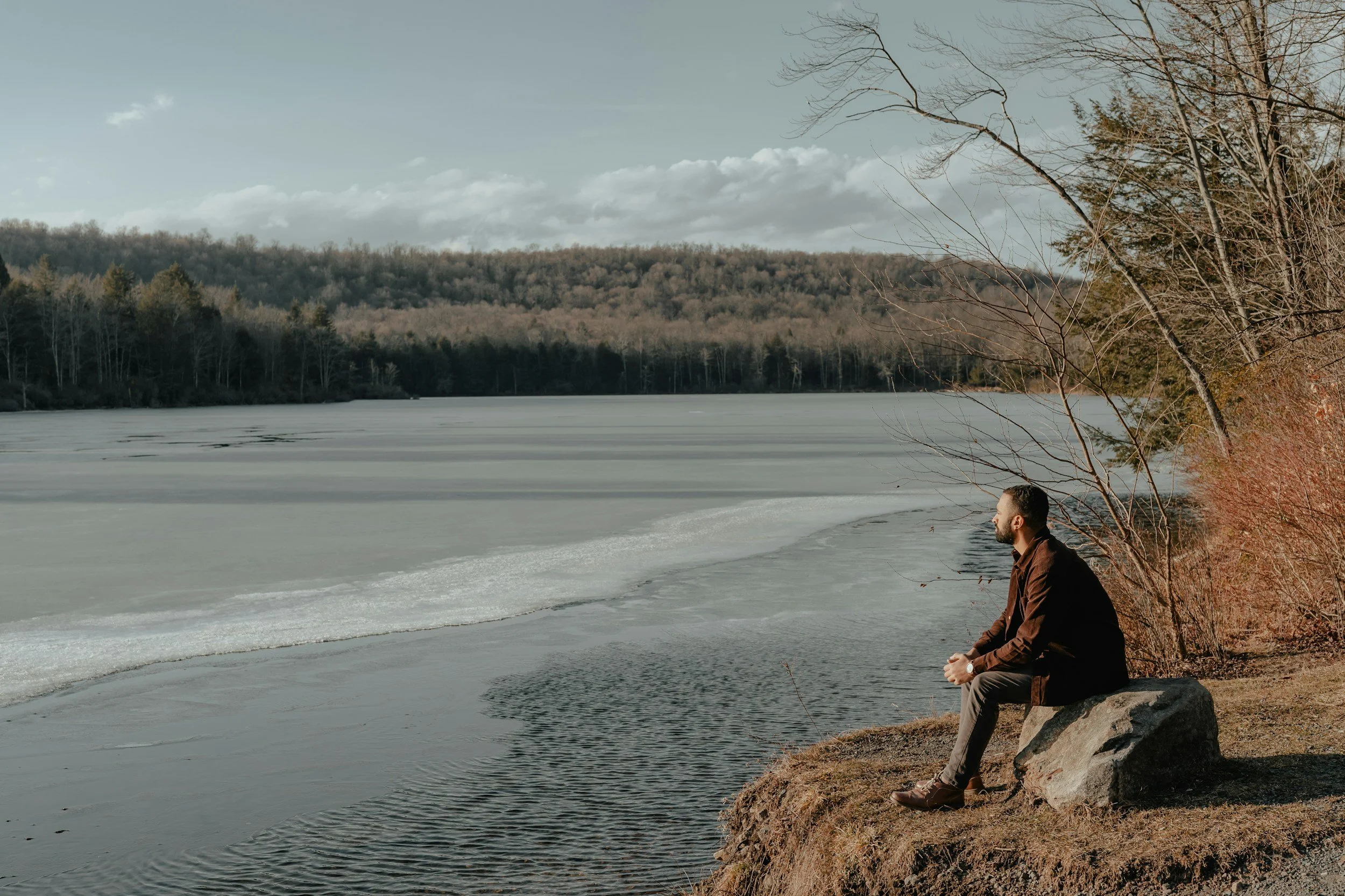 Man sitting on rocks by the edge of a frozen lake, with trees and hills in the background, on a cloudy winter day.