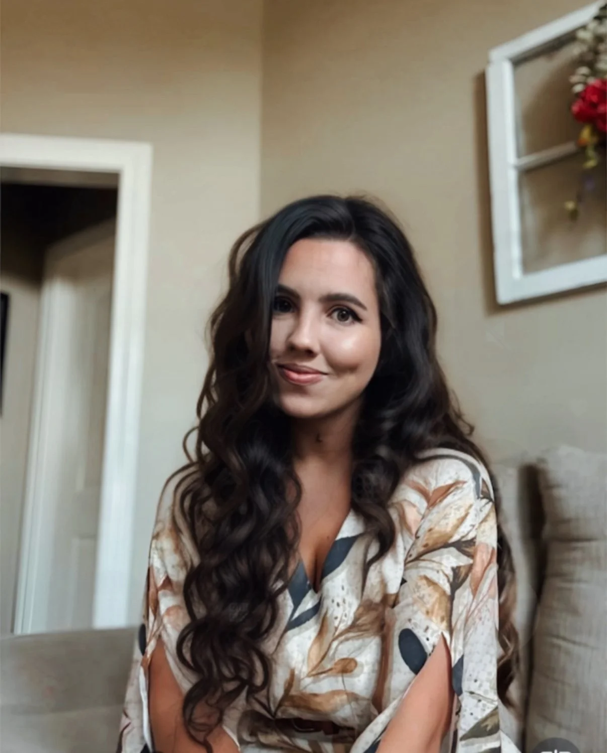 A woman with long, dark, wavy hair smiling while looking at the camera, seated indoors, wearing a patterned blouse, with a beige wall and framed artwork in the background.