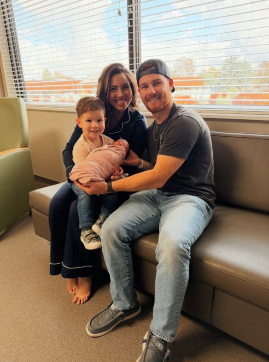 Family of four sitting on a hospital waiting area couch, holding a newborn baby wrapped in a pink blanket, with a large window showing an overcast sky outside.