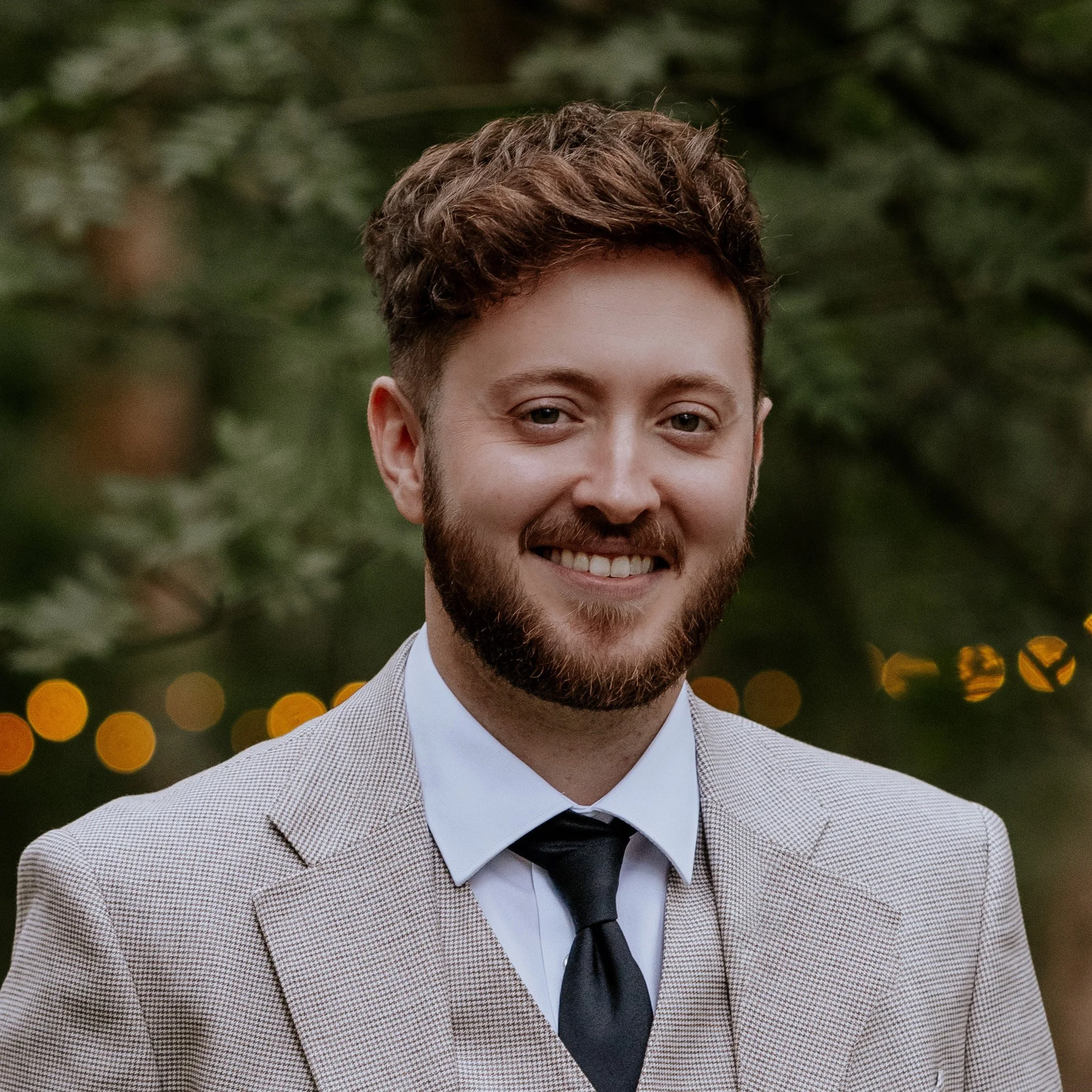 A young man with a beard and wavy brown hair smiling outdoors, dressed in a beige checkered suit, white shirt, and black tie, with a blurred background of greenery and bokeh lights.