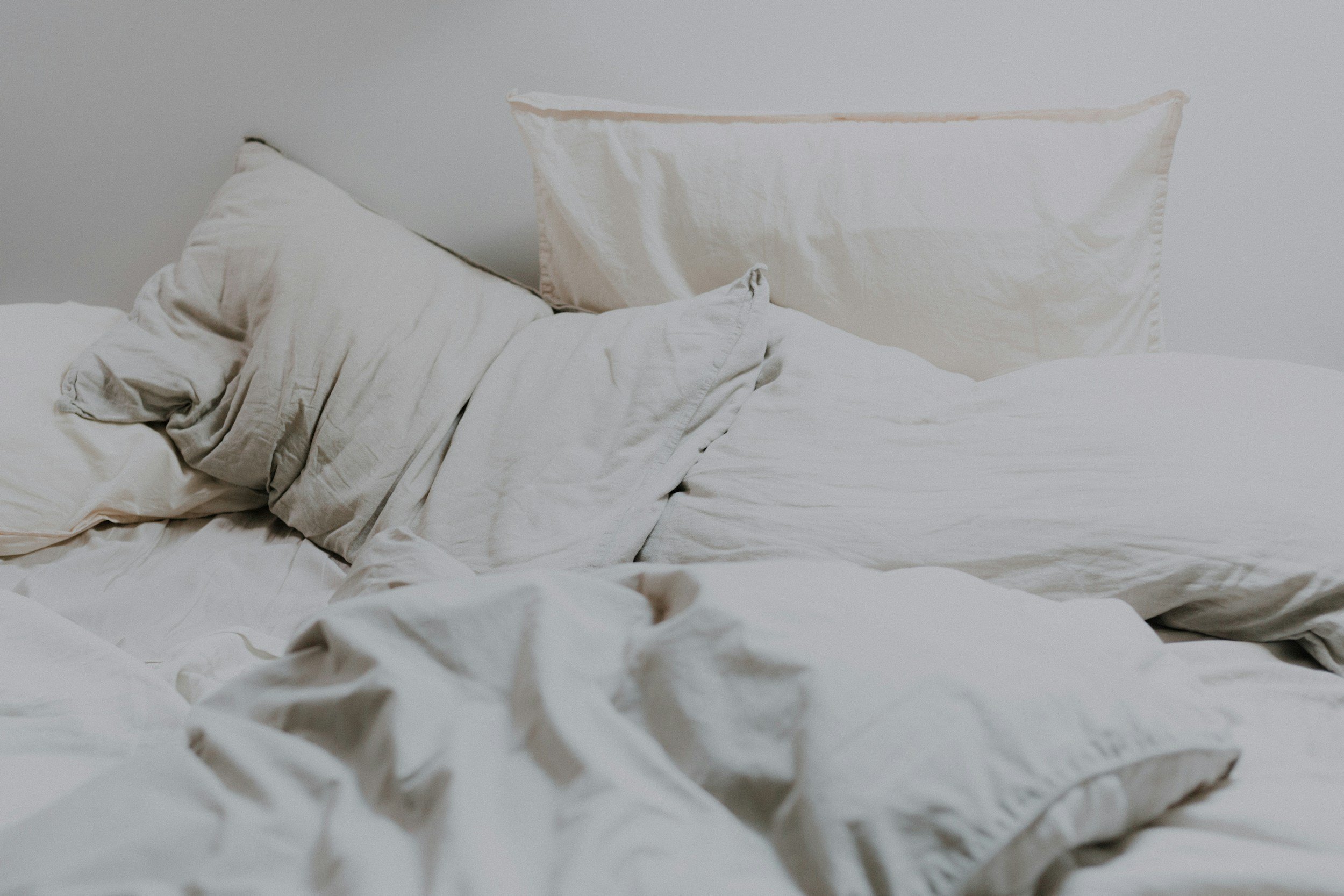 Disheveled bed with crumpled white sheets, pillowcases, and a pillow against a plain wall.