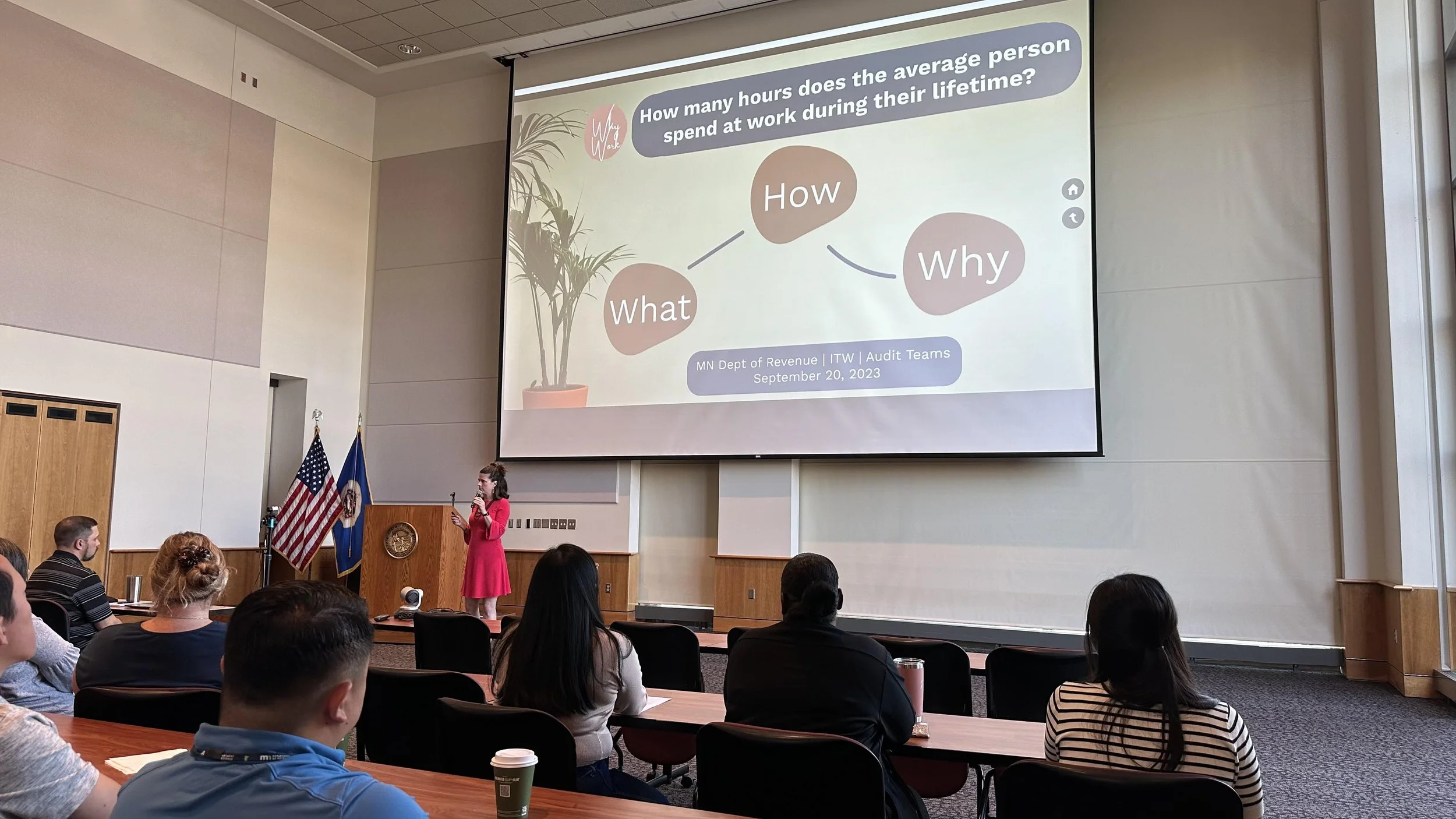 A woman in a pink dress presenting in front of an audience during a conference in a large room with wooden paneling and flags. The presentation slide behind her displays a diagram explaining the question, 'How many hours does the average person spend at work during their lifetime?' with the words 'What,' 'How,' and 'Why.'