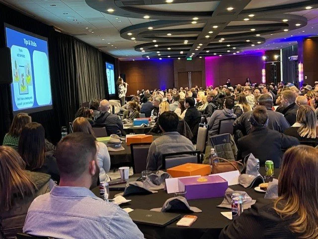 Conference room filled with attendees sitting at tables, watching a presentation on a large screen. The room has a modern ceiling with circular lighting, purple accent lighting at the back, and a presenter standing near the screen.
