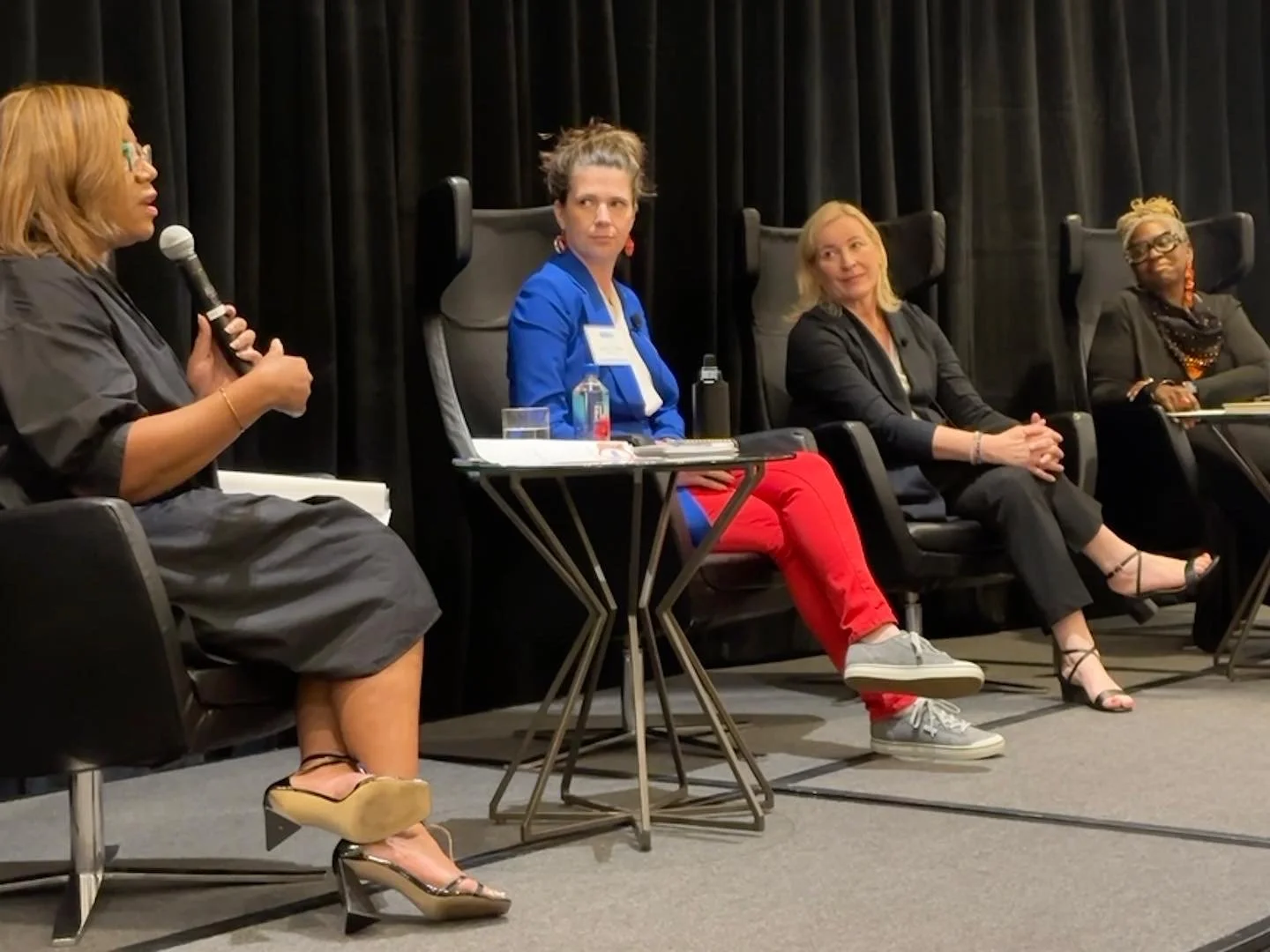 Panel discussion with four women seated on stage, one woman is speaking into a microphone. The stage has a black curtain backdrop.