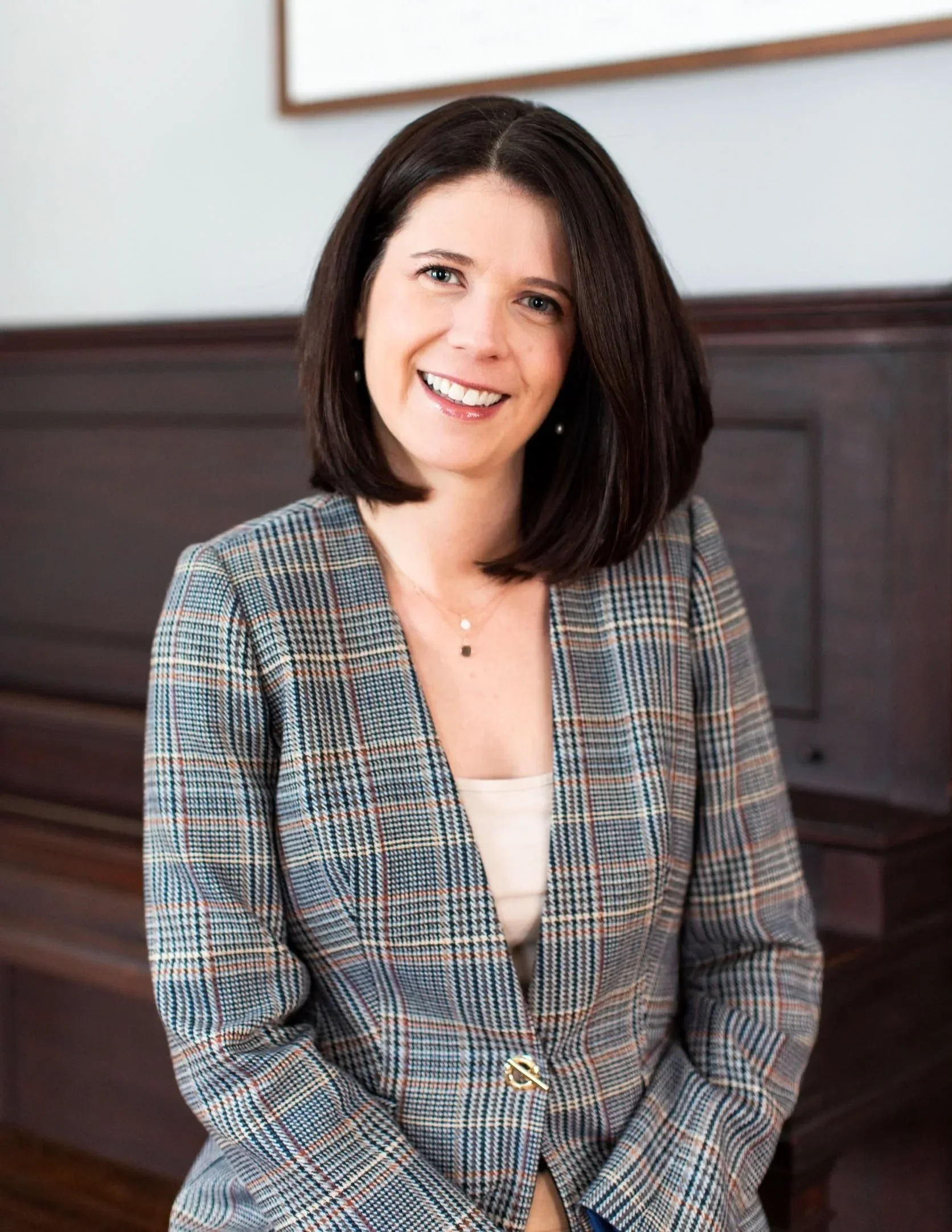 A woman with shoulder-length dark hair smiling and wearing a plaid blazer and a light-colored top, sitting in front of a wooden piano.