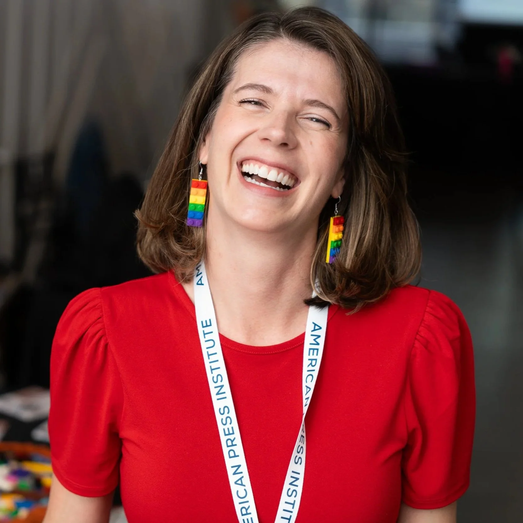 A woman wearing rainbow pride earrings and a red blouse, smiling brightly at an indoor event, with a conference badge hanging around her neck.