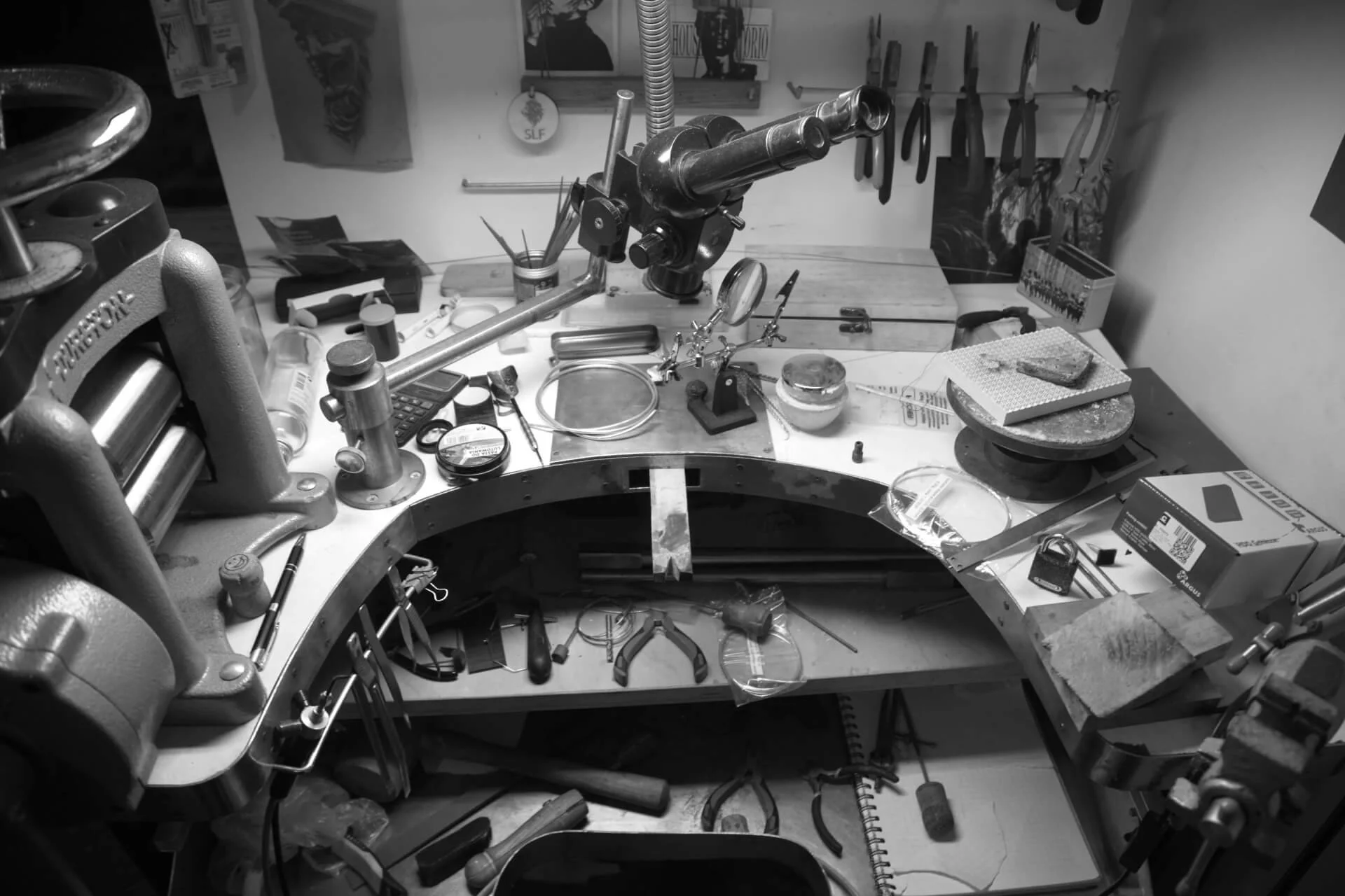 A cluttered workbench in a workshop with various tools, equipment, and materials including a microscope, pliers, a soldering iron, and pieces of stone or metal, surrounding a semi-circular metal frame.