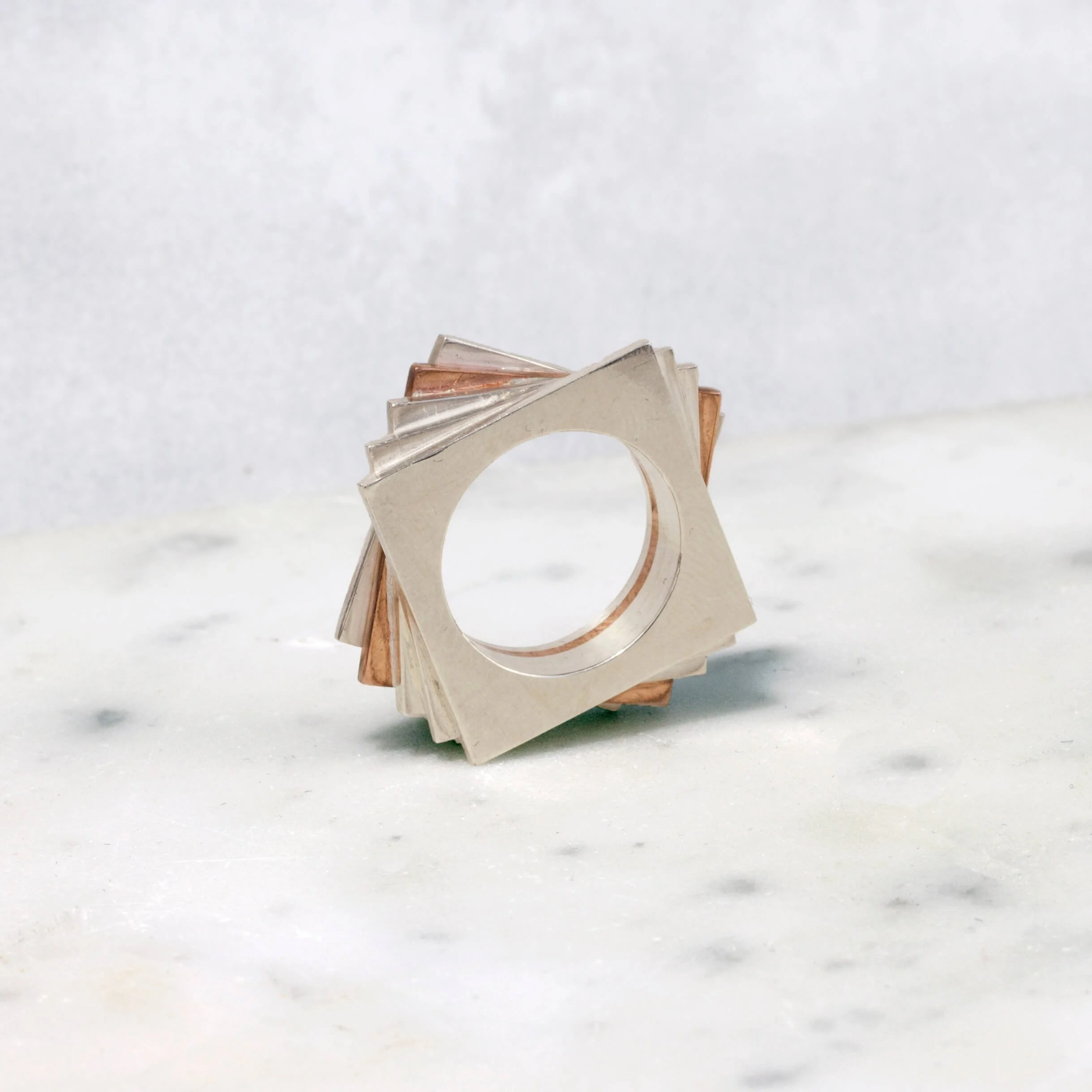 A geometric sculpture made of metal pieces in a layered, square formation with a central circular hole, placed on a white marble surface.