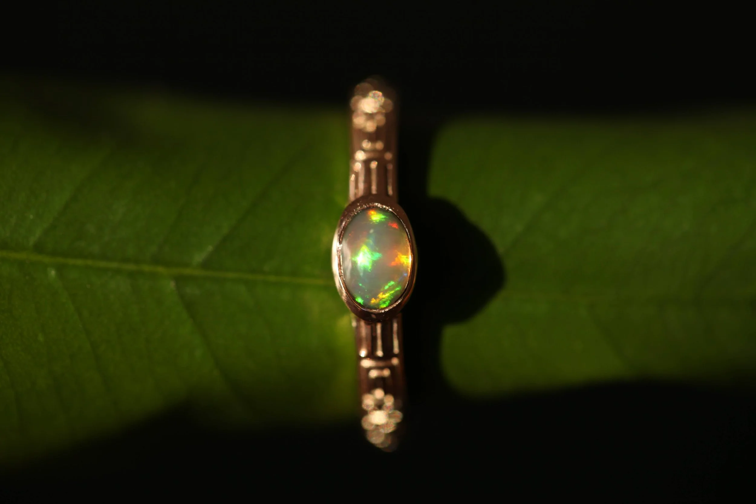 A ring with an oval opal gemstone set in a metal band, placed on a green leaf with a dark background.