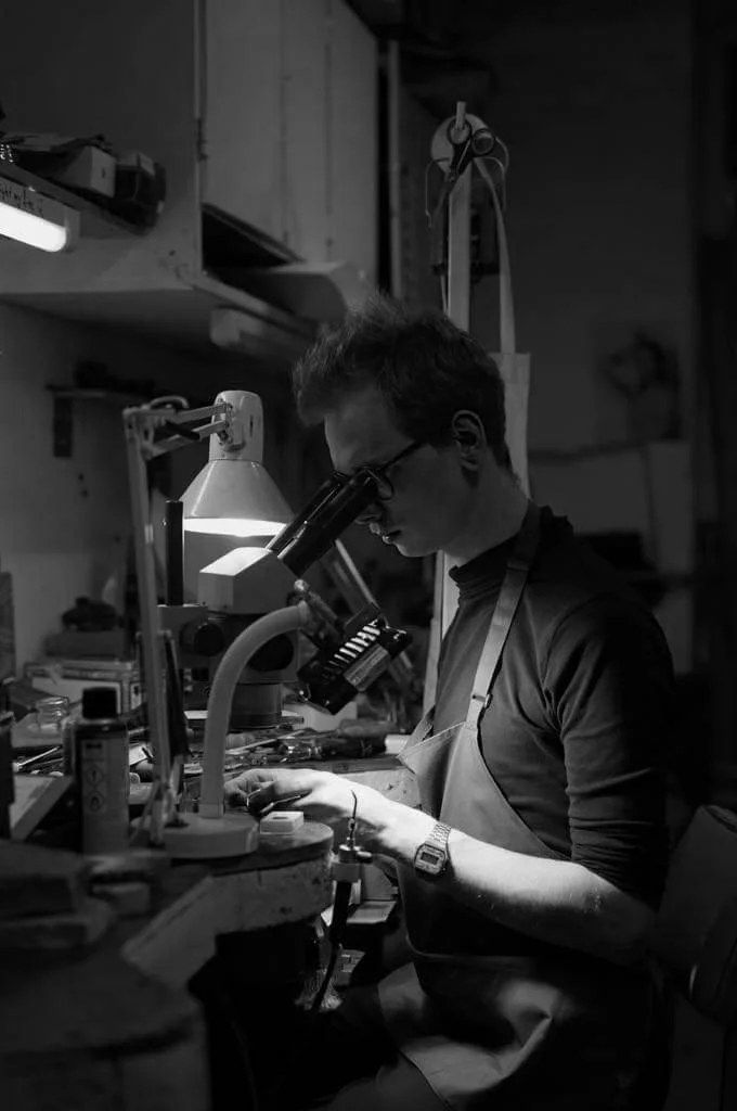 A person working at a cluttered workbench in a workshop, using a microscope and surrounded by tools and equipment, illuminated by a desk lamp.